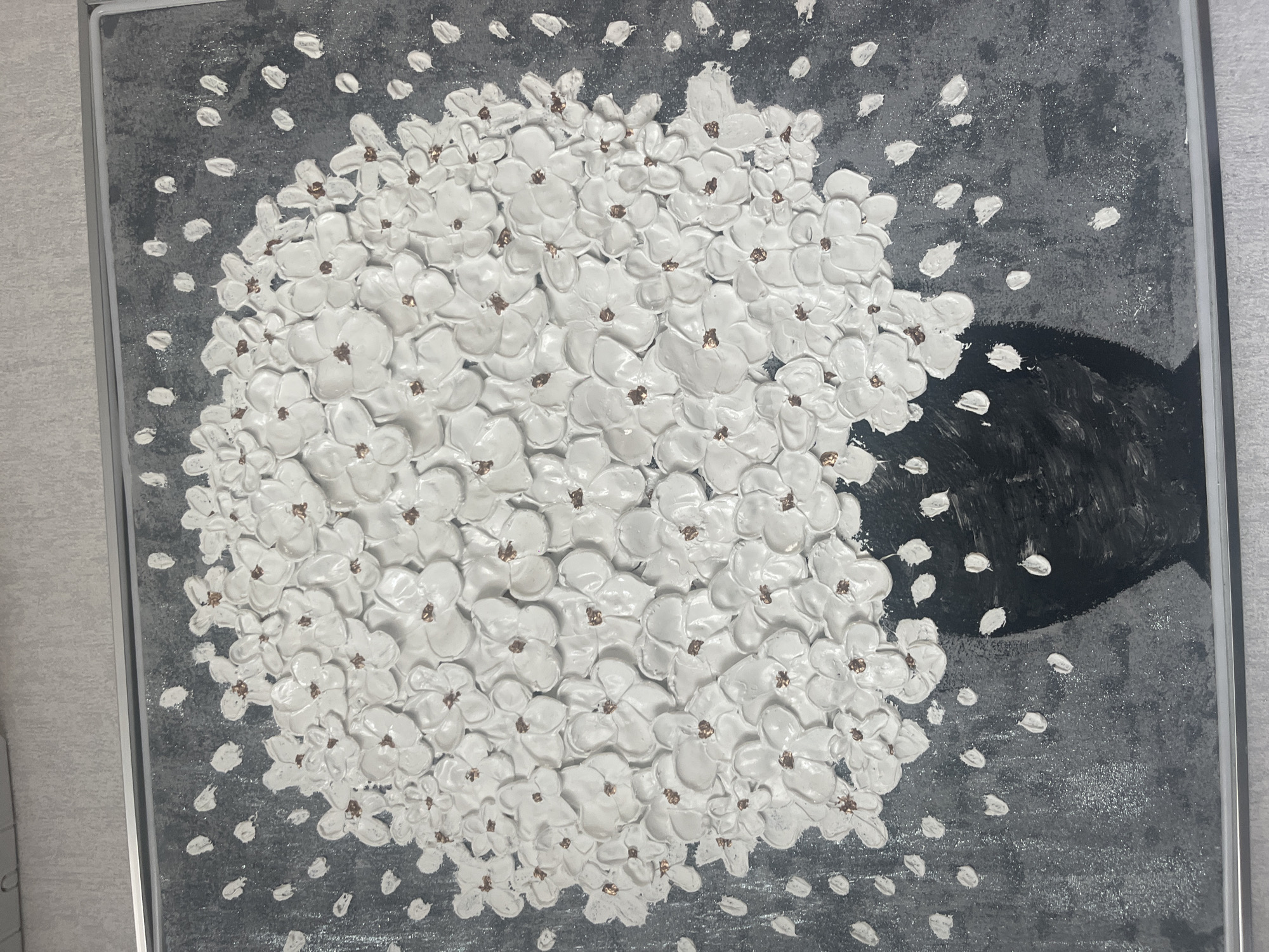 This is a photograph of a textured painting of white flowers in a black vase. The painting is framed in a thin, silver frame. The background of the painting is a dark, textured gray with scattered white dots that appear to be falling petals or blossoms. The flowers are highly textured and three-dimensional, with individual petals clearly defined. The centers of the flowers are a coppery brown color. The vase is depicted as a solid black shape at the bottom of the painting. The photograph is taken from a slightly overhead angle, and the painting is hanging on a light-colored, textured wall. There is no indication of the city of Tyumen, Russia, in the image.