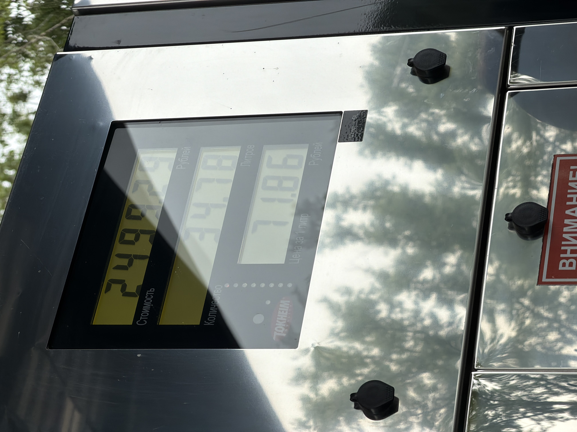 A close-up, eye-level shot captures the digital display and reflective metallic casing of a Tokheim gas pump at a fueling station in Chaikovsky, Russia.
The central feature is an illuminated digital display, presenting three rows of information in yellow segments against a dark background. The top row, labeled "Стоимость" (Cost) in Cyrillic, displays "249.79 Рублей" (Rubles). The middle row, labeled "Количество" (Quantity), shows "34.78 Литров" (Liters). The bottom row, labeled "Цена за 1 литр" (Price per 1 liter), indicates "71.86 Рублей." The "TOKHEIM QUALITY" logo is visible on the lower left of the display screen.
The pump's exterior casing consists of highly polished, reflective metallic panels (likely stainless steel), framed by a darker, possibly black or dark grey, material around the display. This reflective surface prominently mirrors dense green tree foliage and a bright, possibly overcast sky, indicating an outdoor setting with surrounding vegetation. Three black, rubberized caps or plugs are affixed to the lower reflective panels of the pump. A small rectangular sticker with "Vmin = 5L" and other faint text is visible below the display on the right side. Partially visible at the very bottom edge of the image is a red sign with white Cyrillic text, starting "ВНИМАНИЕ!" (ATTENTION!).
No people or explicit actions are depicted.