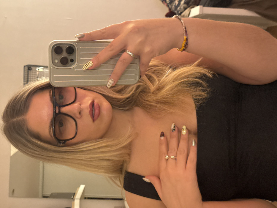 A woman in a black tank top is angled in a selfie, holding her phone with a ribbed silver case. She has blonde hair and wears black glasses. Her nails are decorated with various patterns: polka dots, stripes, and rhinestones. She wears two rings on her right hand and a beaded bracelet on her right wrist. The background includes a mirror reflecting parts of a room.