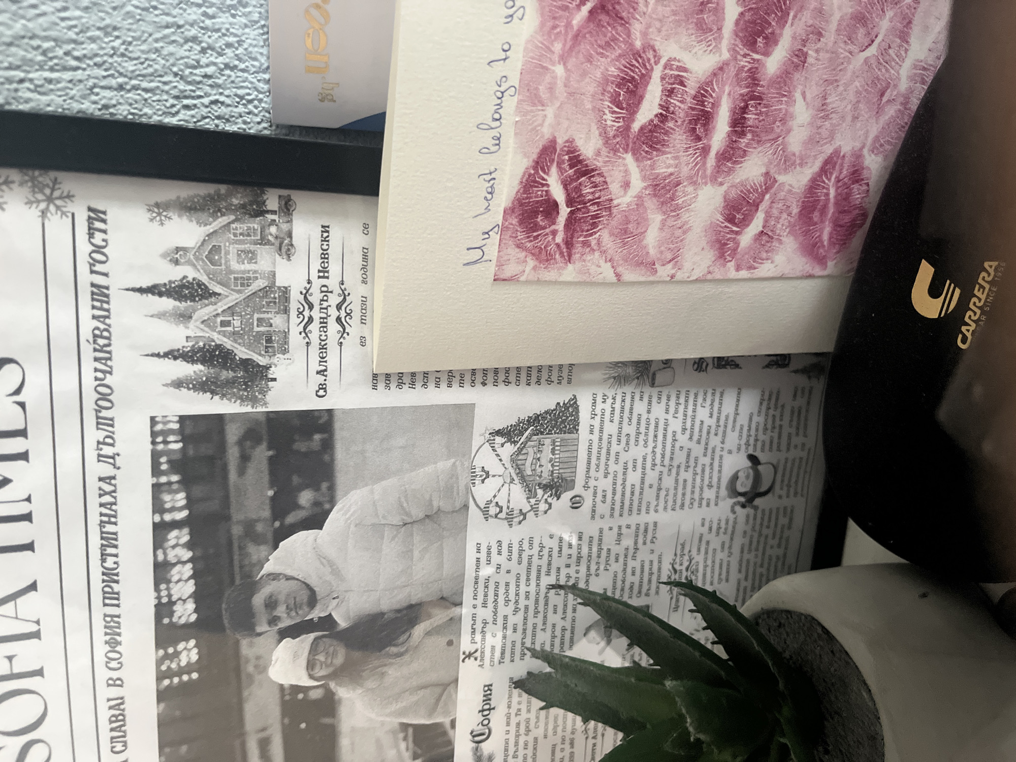 The image displays a rotated, overhead view of various items arranged on a surface, likely a desk, within a Bulgarian context, as suggested by visible text related to Sofia.
The primary subjects include several pieces of printed material: newspaper pages, a greeting card, and a partially visible label or business card. A black object, possibly a case or bag, is visible on the right, along with a small potted succulent plant in a white pot towards the bottom.
The activity captured is a static arrangement of these items, suggesting a personal space or collection of mementos.
Notable details include:
- A greeting card with a cream border featuring numerous pink lipstick kiss marks and the handwritten message "My heart belongs to you," indicating a theme of affection.
- Newspaper clippings, predominantly in Bulgarian, featuring black and white images. One image shows a couple, a man and a woman, dressed in winter coats and hats, suggesting a cold season. Another image depicts a snow-covered house amidst pine trees, and a Ferris wheel, further reinforcing a winter or holiday theme.
- The black object bears the "CARRERA" logo and the text "YEAR SINCE 1956."
- A partially visible label shows "reen.bg".
Visible text on the newspaper clippings includes "SOFIA TIMES" (partially visible), "СЛАВАЇ В СОФИЯ ПРИСТИГНАХА ДЪЛГООЧАКВАНИ ГОСТИ" (GLORY IN SOFIA, THE LONG-AWAITED GUESTS ARRIVED), and "Св. Александър Невски" (St. Alexander Nevsky). Other Bulgarian text mentions "София" (Sofia), "България" (Bulgaria), "Александър Невски" (Alexander Nevsky), and historical references such as the Teutonic Order, Lake Peipus, and the First World War. The content of the newspaper appears to discuss historical or cultural events related to Sofia and Bulgaria, aligning with the general location context of Pokrovnik, Bulgaria.