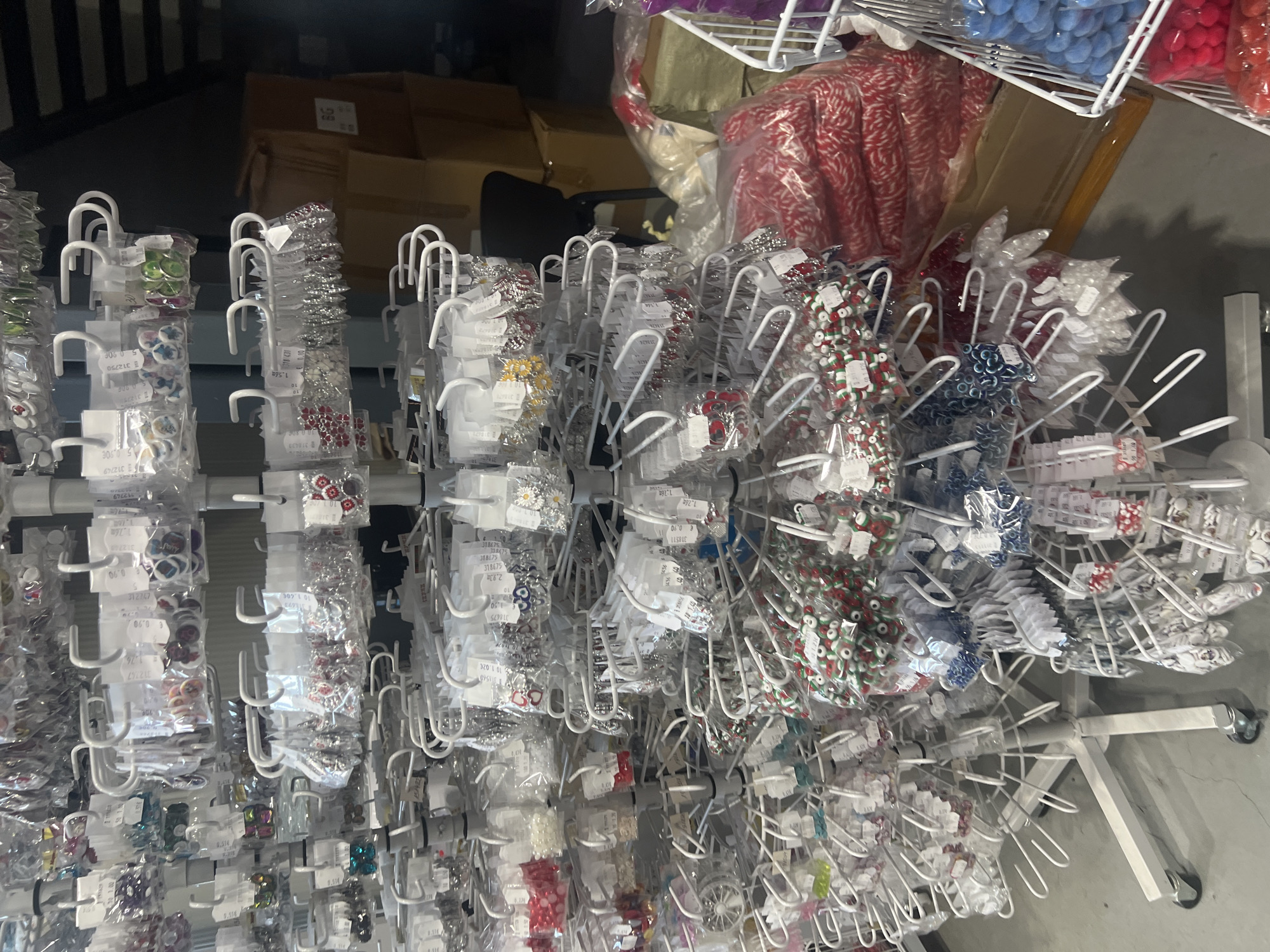 The image shows a retail display of small, individually packaged craft supplies, likely beads and decorative elements, hanging from a series of white metal racks. The racks appear to be part of a rotating display stand on wheels, which is positioned on a gray floor. In the background, there are boxes and possibly a chair, indicating a storage or back-of-house area. There are no people visible. The location is potentially a craft store or a department store with a craft section, possibly located in Burgas, Bulgaria, due to the product labeling using "31" as part of their item number.