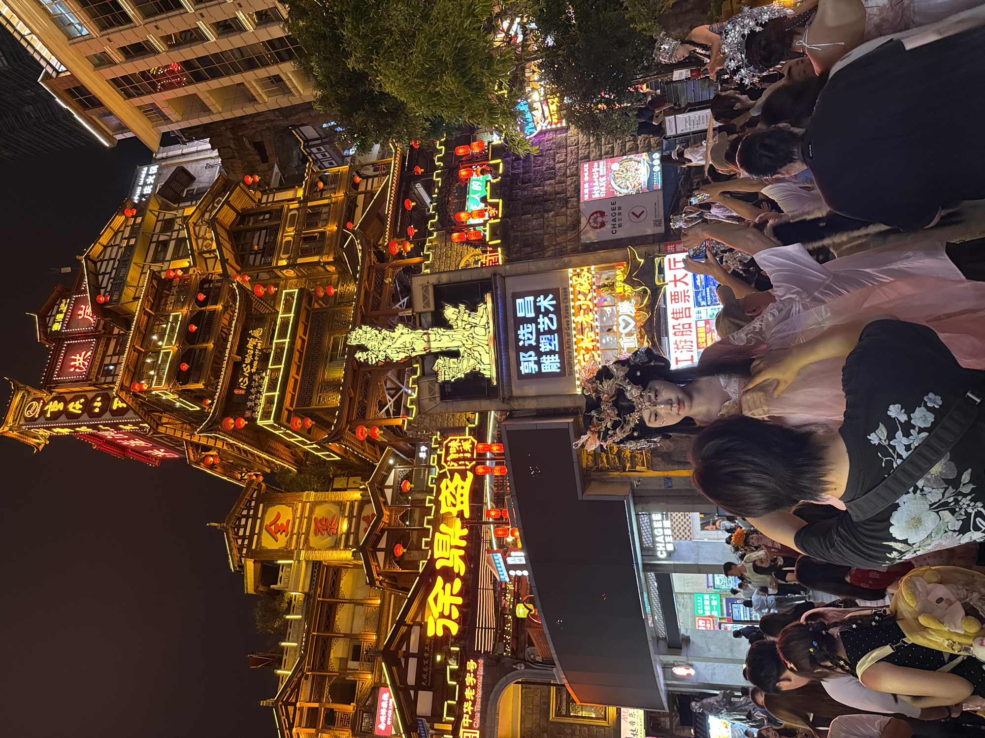 This is a night-time outdoor shot in Yuzhong District, China, focusing on a bustling pedestrian street. The dominant feature is a tall, multi-story building with traditional Chinese architecture, illuminated by numerous red lanterns and neon signs, some of which display Chinese characters. The building is ornately decorated with multiple tiers and intricate wooden structures.
In the foreground, a crowd of people is gathered. A young woman, dressed in a detailed traditional Chinese costume with an elaborate headpiece, stands as a focal point. She is adorned with makeup, and her hair is styled elaborately. Several people in the crowd are taking photographs of her with their mobile phones. The crowd appears diverse, with individuals of various ages and styles of dress, some in modern attire and others possibly in costumes themselves.
The environment is urban, with other buildings visible in the background, some of which are modern high-rises. There are trees and street lights contributing to the overall ambiance. The ground appears to be paved. The overall scene conveys a sense of festivity and cultural display.