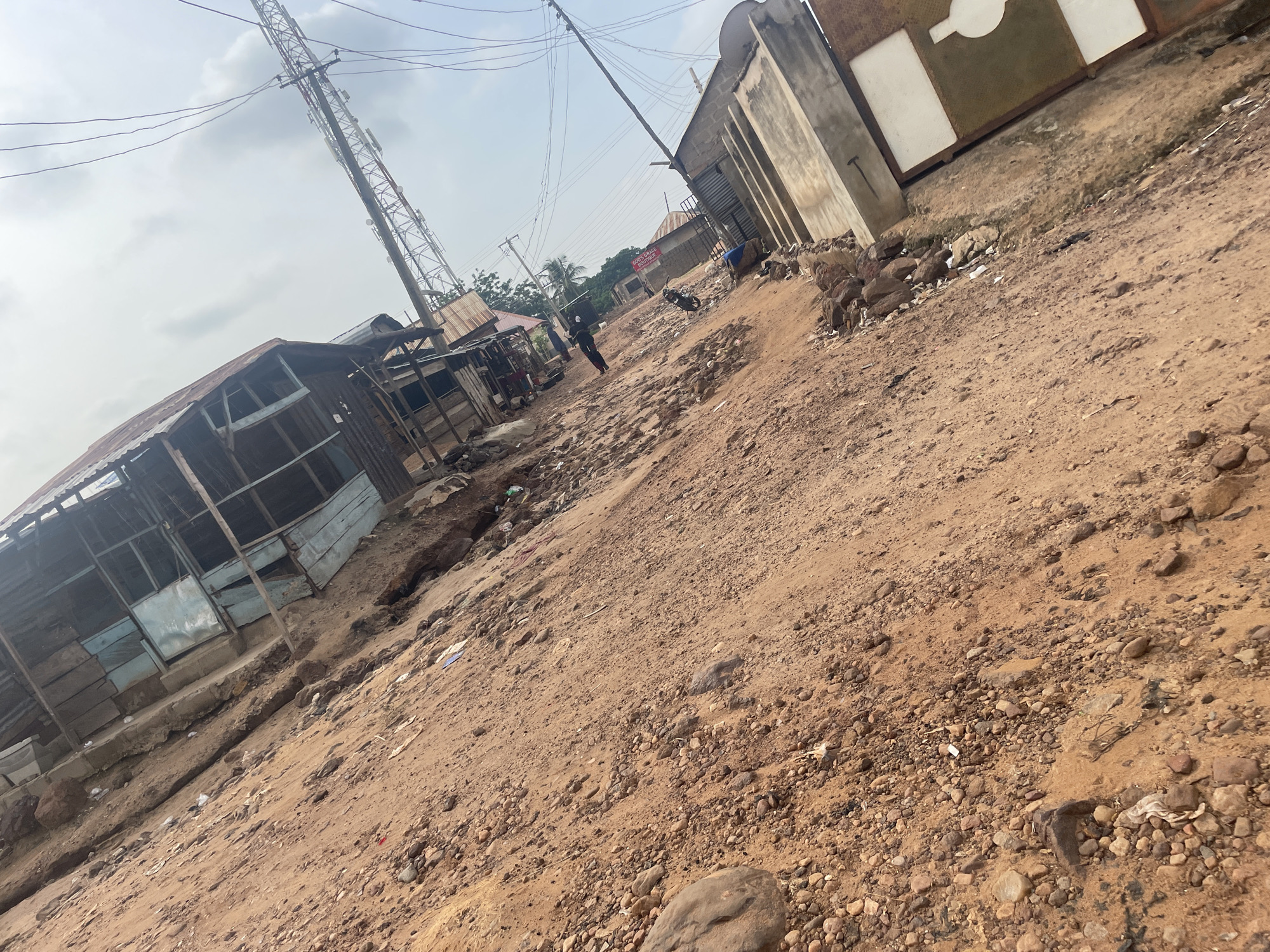 The image depicts a tilted, overhead view of a dirt road in Lokoja, Nigeria, with informal market stalls lining one side and a concrete structure on the other. The market stalls appear to be constructed from wood and corrugated metal, some in disrepair. Scattered debris, including plastic waste and rocks, litters the dirt road. In the distance, a telecommunications tower and power lines are visible, along with palm trees suggesting a tropical climate. A motorcycle is parked on the side of the road, and a few individuals, whose features are indistinct, are present in the background. The sky is overcast with clouds.