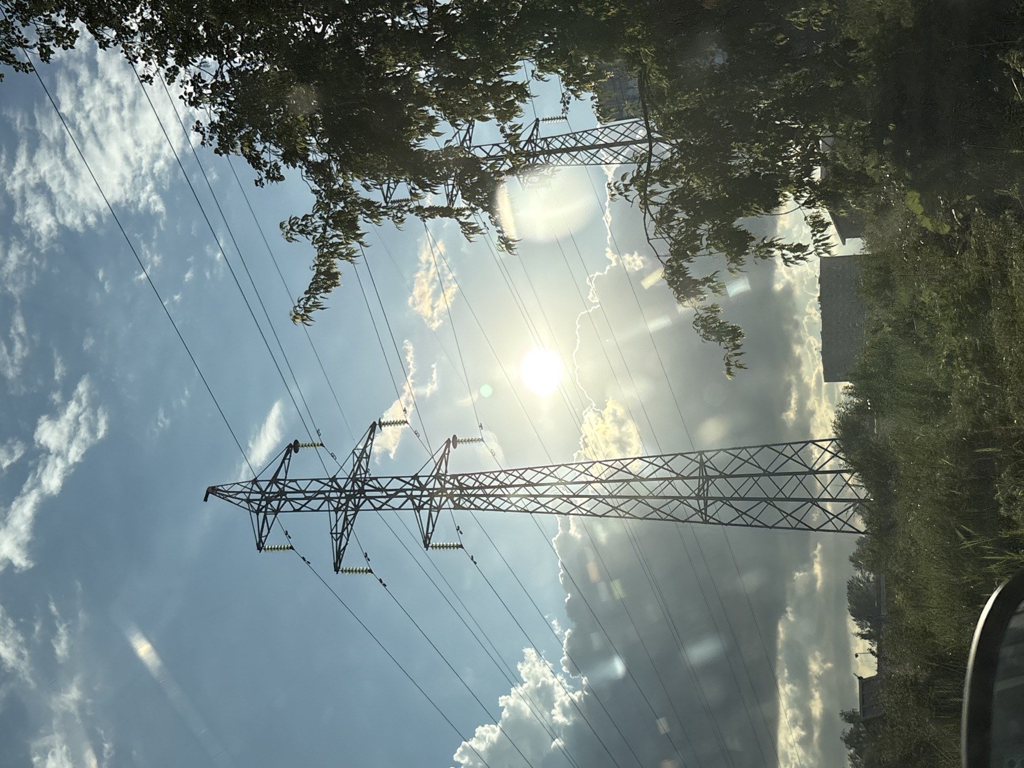 The image depicts a power line tower standing tall amidst a sunlit sky with scattered clouds, a common sight in the outskirts of Saint Petersburg, Russia. The tower, made of steel lattice, supports multiple high-voltage wires that stretch across the frame. To the right, a dense canopy of green leaves from a tree partially obscures the tower and the sky, creating a natural contrast with the industrial structure. The sun is visible, partially obscured by clouds, casting a bright glow and creating lens flares. In the background, a low-rise building can be seen, adding to the suburban or industrial landscape characteristic of areas surrounding large cities like Saint Petersburg. The foreground shows lush green vegetation and the edge of a vehicle, suggesting the photo was taken from a moving car. The scene is set during daytime, with the sunlight indicating it is likely afternoon or late morning.