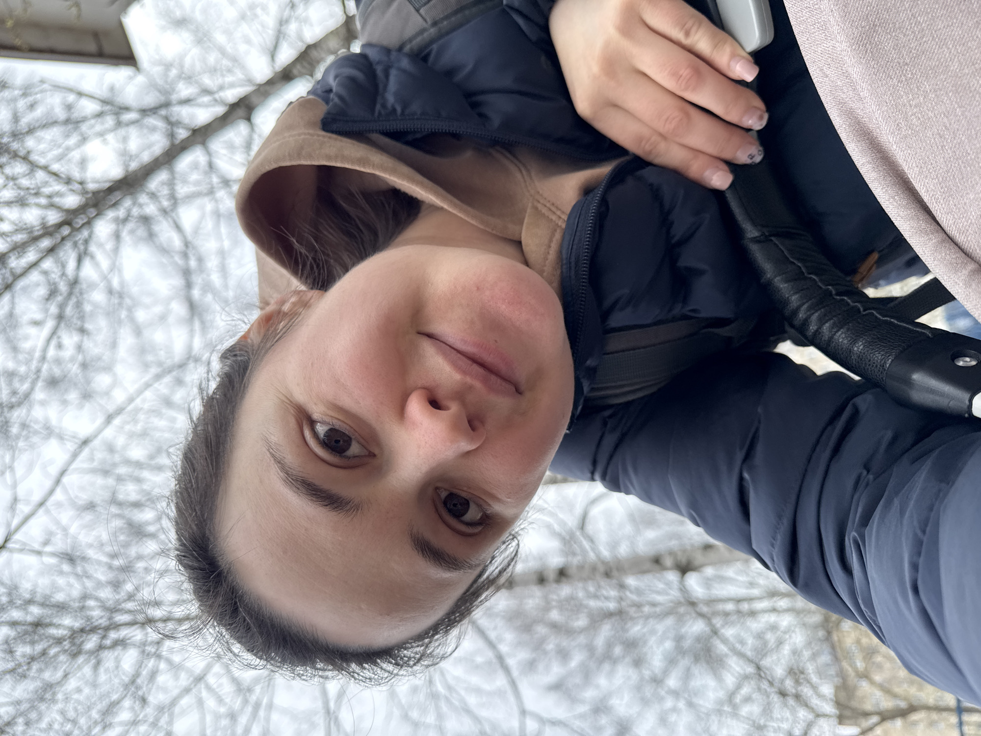 A selfie of a woman in Izhevsk, Russia, taken outdoors in winter. She is wearing a beige hoodie underneath a dark blue jacket. She is holding the handlebar of a baby stroller. The background shows bare tree branches against a gray sky, suggestive of a park or wooded area. The woman is fair-skinned with brown eyes and dark hair pulled back from her face. Her nails are painted light pink with designs on some. The angle is slightly downward, indicating she is holding the camera above her.