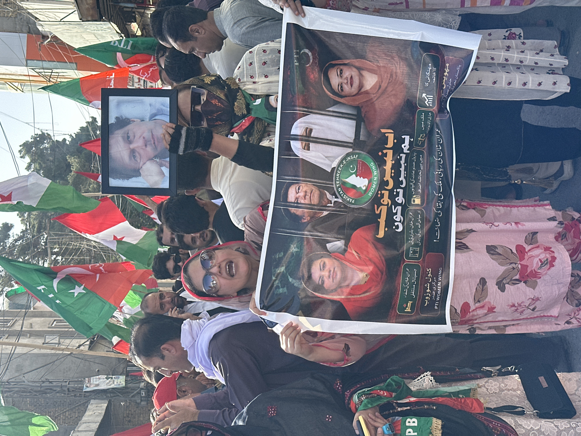 A political protest is taking place in what appears to be Quetta City Tehsil, Pakistan. People are gathered holding a variety of signs and flags. A framed portrait of a man is being held aloft. A large banner with images of multiple people, including the man from the portrait, is also being held. The banner includes text in Urdu and the logo for "Pakistan Tehreek-e-Insaf Women Wing". The protestors are diverse in age and gender. Some are wearing sunglasses and traditional clothing.