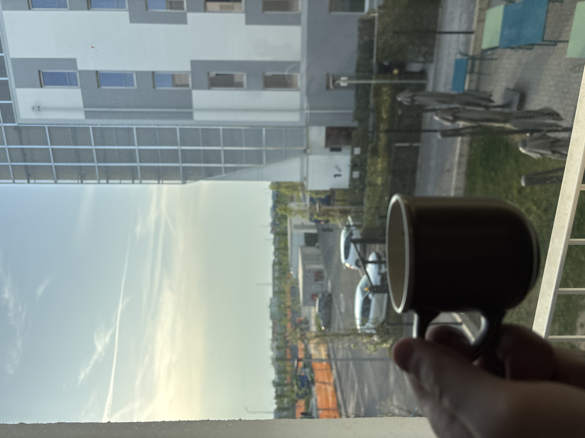 The photo depicts a view through a window, with a hand holding a dark-colored mug in the foreground. The window frame is visible at the bottom of the frame. Outside, a multi-story building with rows of windows is partially visible, as well as trees and vehicles parked along a street. The sky is pale blue with wispy clouds. The city is Novate Milanese, Italy.