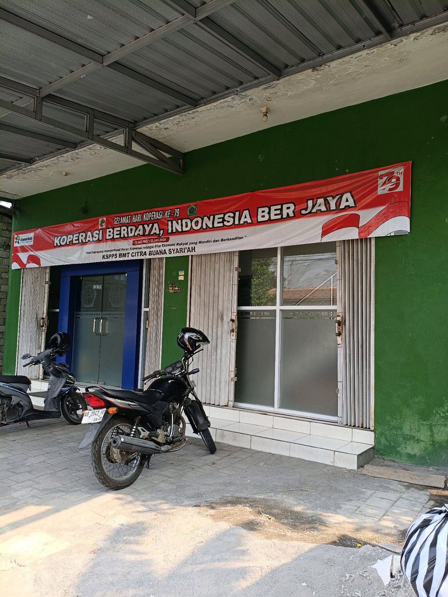 In front of a green-painted building in Maguwoharjo, Indonesia, a red and white banner reading "SELAMAT HARI KOPERASI KE-79. KOPERASI BERDAYA, INDONESIA BER JAYA" is displayed above a glass-fronted entrance. The banner also includes text in smaller print stating "12 JULI 1947 - 12 JULI 2026" and "Pilar bersama memperkuat Peran Koperasi sebagai Pilar Ekonomi Rakyat yang Mandiri dan Berkeadilan". Below the banner, two motorcycles are parked on a paved area. The motorcycle on the left is partially visible and appears to be a scooter. The motorcycle on the right is a black motorcycle with a license plate that reads "AB 2942 AD". The entrance has blue-framed glass doors with "BUKA" (Open) written on them and frosted glass panels. To the right of the entrance are metal accordion doors. The building has a metal roof supported by visible metal framing. The ground in front of the building is paved with concrete tiles and there are some piles of sand and gravel, possibly indicating construction or repair work. A black and white striped trash bag is visible in the lower right corner of the image. The overall scene depicts a street-level view of a business establishment.