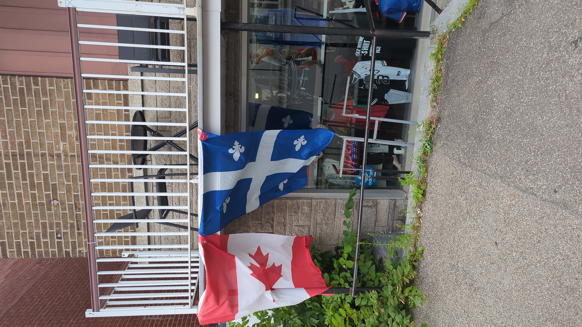 Two flags, the Canadian flag and the Quebec flag, are hanging on the facade of a store. The Canadian flag, on the left, is red and white with a red maple leaf in the center. The Quebec flag, on the right, is blue with a white cross and four white fleurs-de-lis. Both flags are attached to a white railing that runs along the top of the store's storefront. Behind the railing, a small balcony area is visible, with a table and two chairs. The store's interior is visible through the glass windows, with clothing items displayed on racks. The store appears to be a clothing boutique. The storefront is made of brick. The image was taken from the sidewalk in front of the store, which is paved with asphalt. The scene is set in Montreal, Canada.