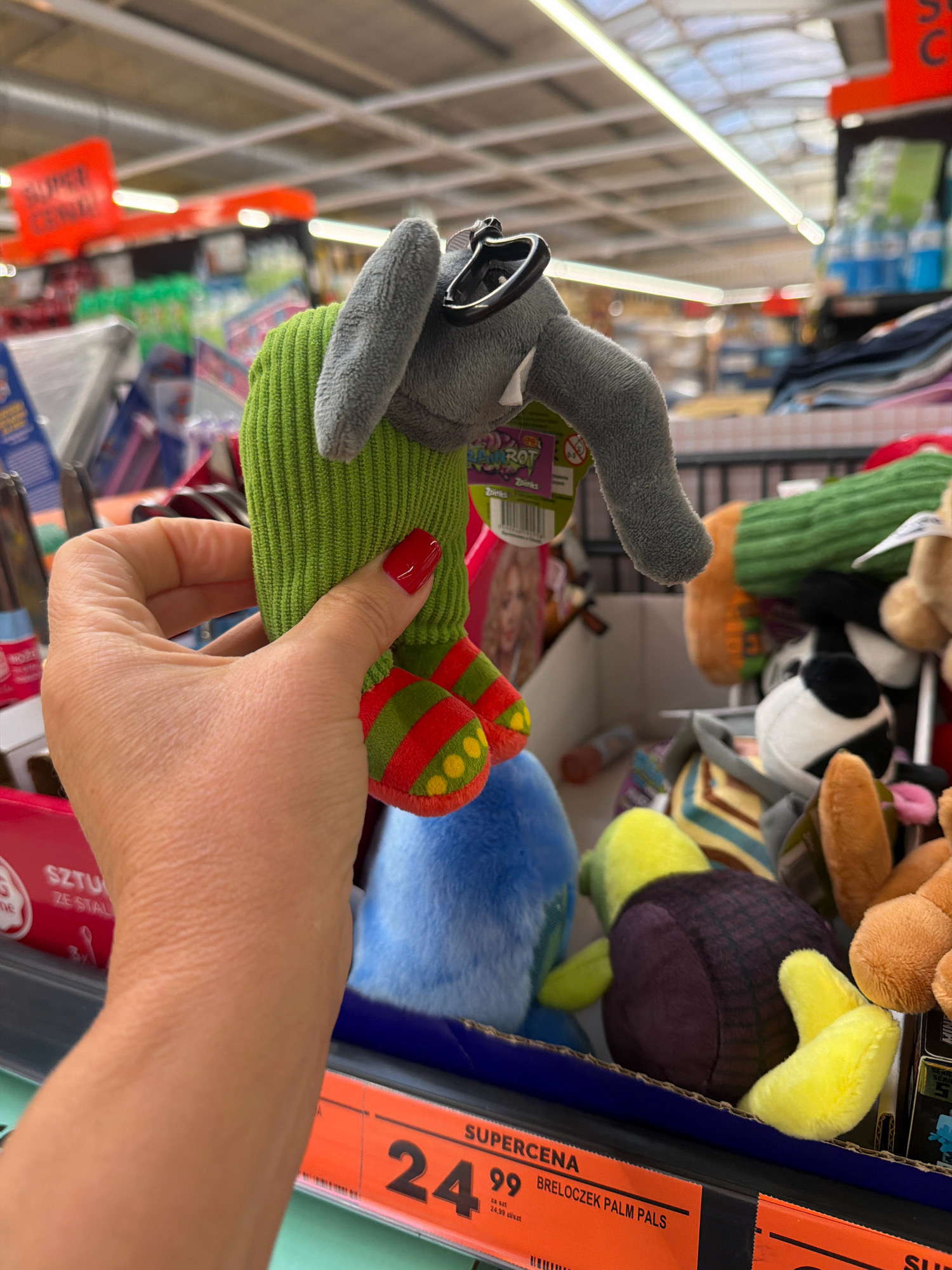 A person's hand with red nail polish is holding a small, grey plush elephant toy with a green ribbed body and red and green striped feet with yellow polka dots. The toy has a black keyring attached to its head. In the background, shelves are filled with various plush toys and other products. There are also price tags visible. The scene appears to be inside a retail store, potentially a supermarket or a toy store, with a price tag indicating "SUPERCENA" (Super Price) and "24.99 zł/szt" (24.99 Polish Zloty per piece), suggesting the location is in Poland. The ceiling has fluorescent lighting and a grid-like structure. Based on the currency and language, the location is likely a retail establishment in a city like Wrocław, Poland.