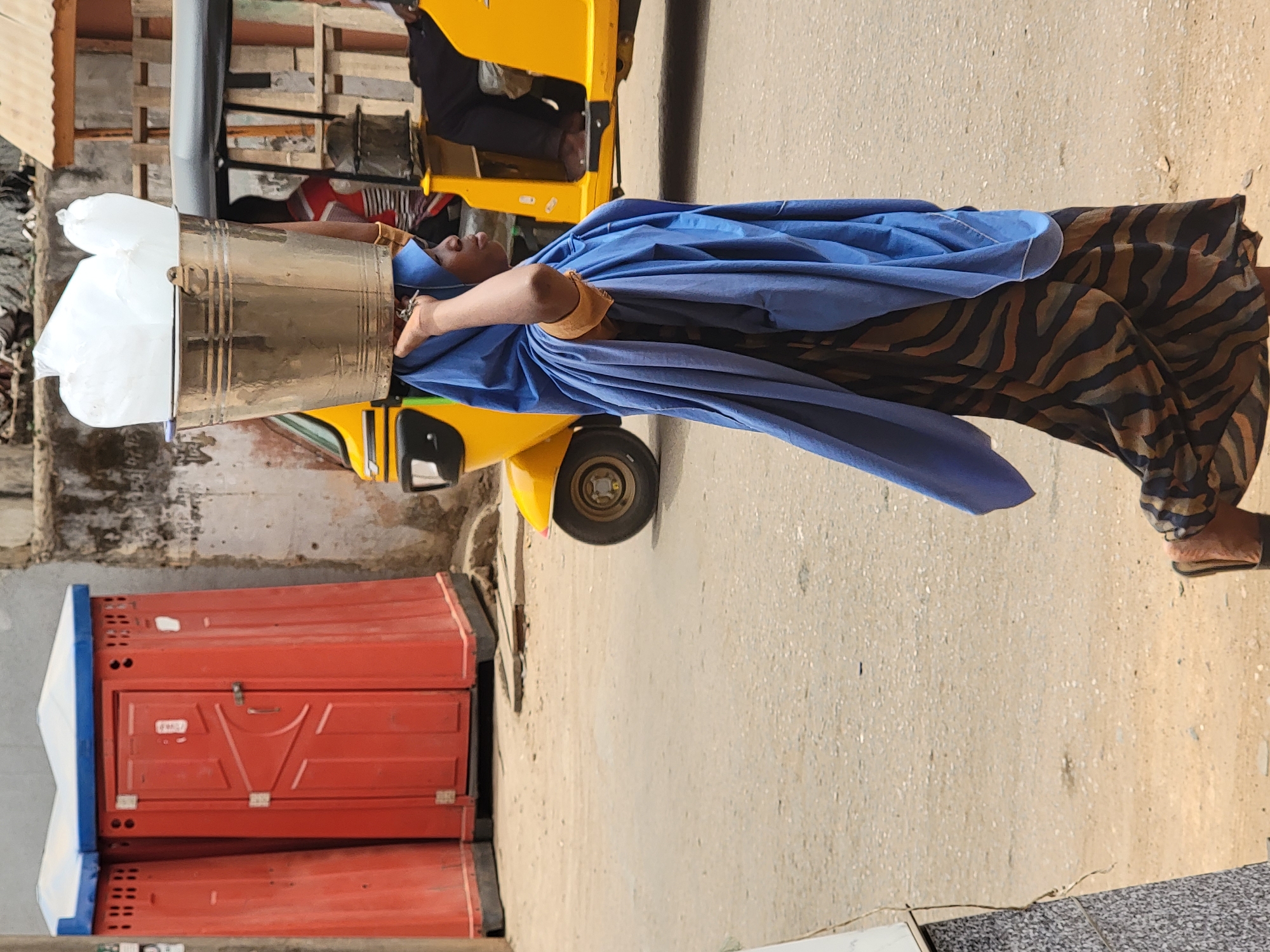 In Maiburiji, Nigeria, a woman carries a silver bucket containing white bags on her head while walking on a concrete street. She wears a blue cloth draped over her head and shoulders, and a patterned brown and black garment. The bucket appears to contain ice. Adjacent to the woman and on the left of the frame is a yellow three-wheeled vehicle. To the left of the vehicle, two stacked orange portable toilets are visible. The ground is a light tan color.
