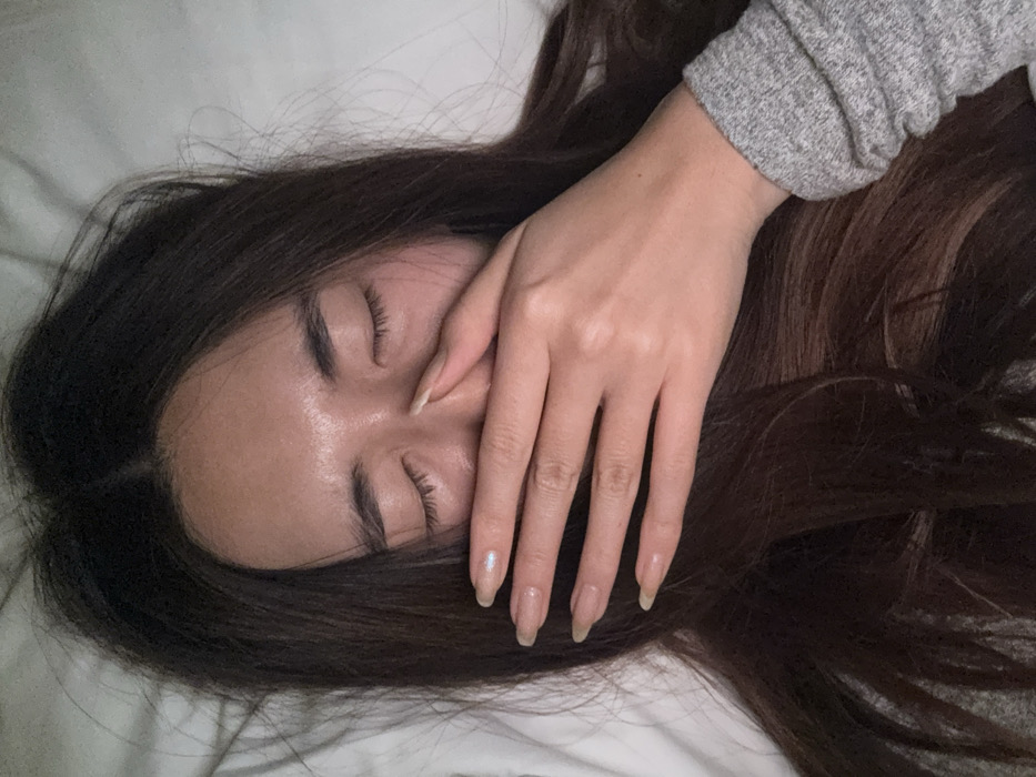 A close-up, top-down view depicts an individual, likely a female, with long dark brown hair, lying down in an indoor setting identified as Le Havre, France. The eyes are closed, and the right hand is positioned over the mouth and lower nose, with the thumb and index finger contacting the bridge of the nose. The individual's skin on the forehead exhibits a slight sheen. Fingernails are long and oval-shaped, with a pale iridescent finish visible on the ring finger. A gray, ribbed long-sleeved garment is visible on the lower right arm. The background consists of white fabric, indicative of bedding, such as a pillow or sheet. The specific location within Le Havre, France, is not visually discernible from the image, which focuses exclusively on the individual and their immediate personal space.