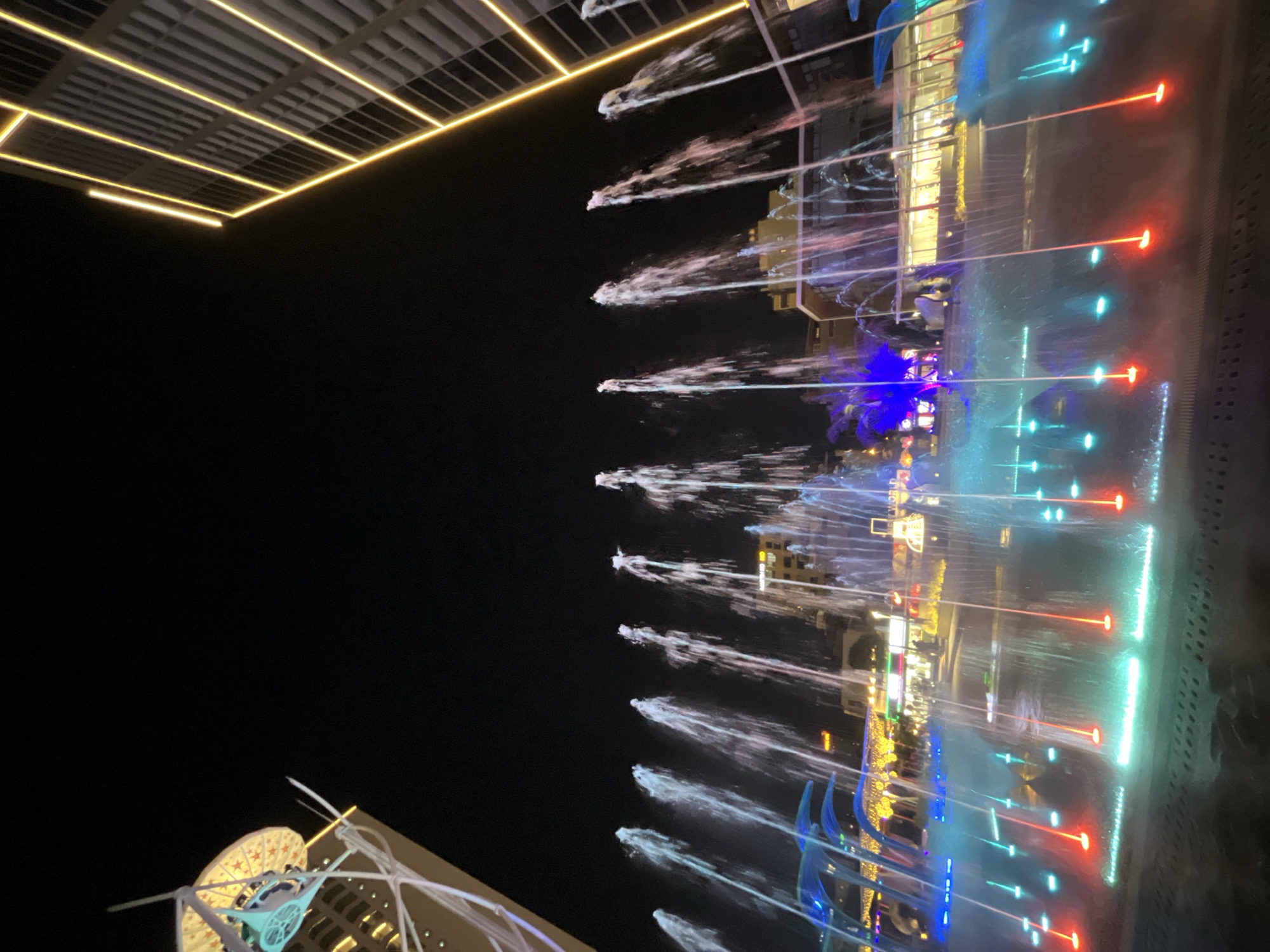 The image shows a vibrant nighttime scene of a modern urban plaza in Da Nang, Vietnam, likely around Dragon Bridge or a similar waterfront area. The foreground is dominated by a large, multi-colored light and water fountain display. Jets of water are illuminated with a spectrum of colors, including bright green, blue, red, and pink, creating a dynamic visual effect as they shoot upwards against the dark night sky. The water forms a shimmering curtain that partially obscures the background.
In the mid-ground, several buildings are visible, illuminated by decorative lighting. One building on the right is a modern structure with prominent yellow and white vertical light strips outlining its facade. Another building on the left, partially visible, features a distinctive architectural element topped with a circular, star-patterned structure. The architecture suggests a well-developed commercial or entertainment district.
The atmosphere is lively and festive, implying a public space where people gather to enjoy the display. Although no specific individuals are clearly discernible due to the distance and lighting, the presence of structures and the nature of the scene suggest a public gathering place. The overall impression is one of a modern, well-lit, and entertainment-focused urban environment.