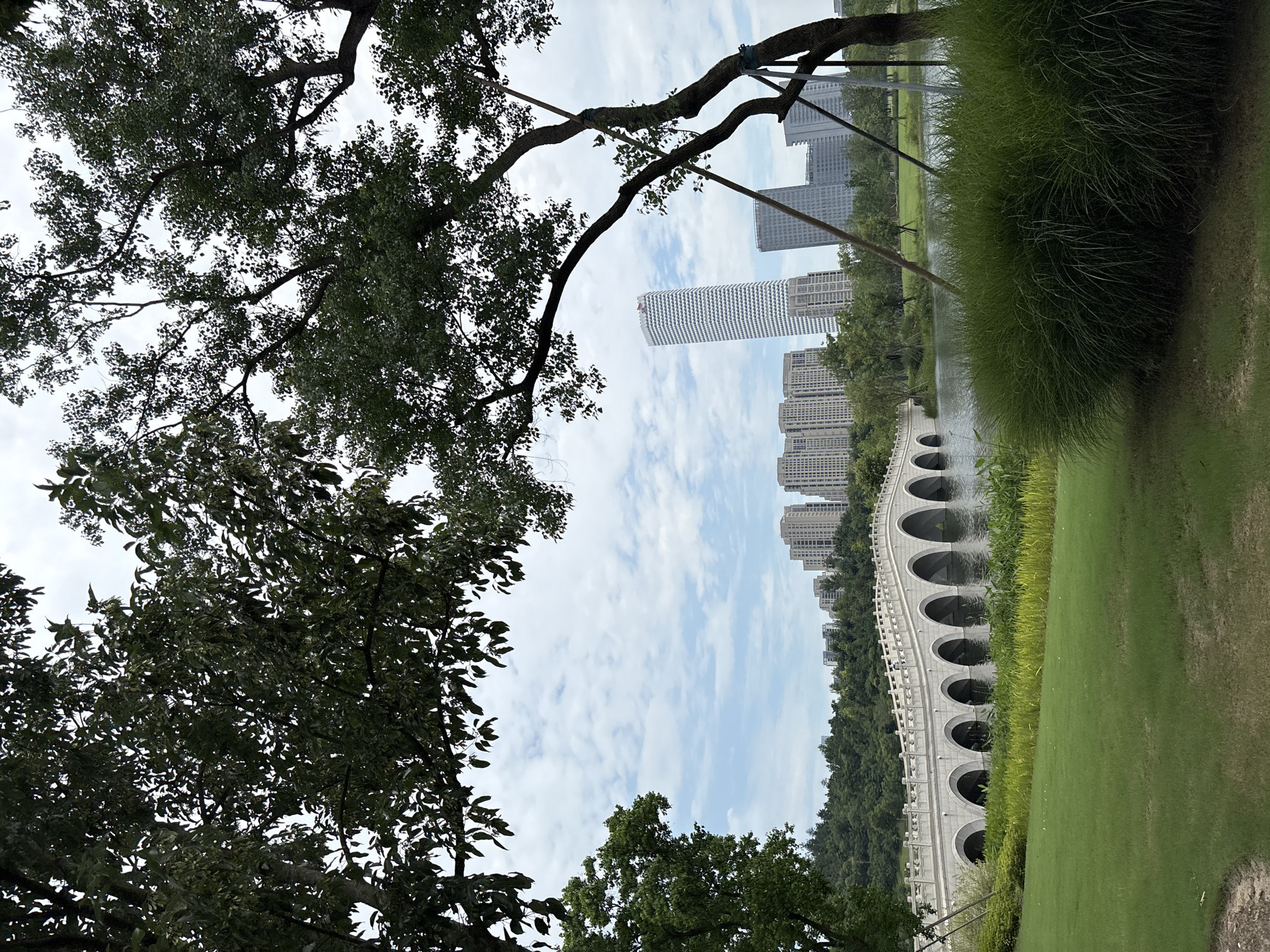 In the city of Pudong, China, a serene park scene unfolds. In the foreground, lush green grass covers the manicured lawn, with a large, bushy clump of ornamental grass to the right. Beyond the grass, a tranquil lake reflects the cloudy sky and surrounding greenery. Spanning the lake is an elegant, arched stone bridge with multiple arches, its white stone structure contrasting with the dark water.
In the background, a densely forested hillside rises behind the lake, dotted with trees. A cluster of modern high-rise buildings, including a distinctive white, cylindrical skyscraper, forms the urban backdrop to this natural oasis. The sky above is partly cloudy, with white clouds scattered across a pale blue expanse. Trees with dark green foliage frame the top and sides of the image, partially obscuring the sky and adding depth to the composition. The scene is peaceful and well-maintained, suggesting a public park or recreational area within the bustling city. No people are visible in the image.