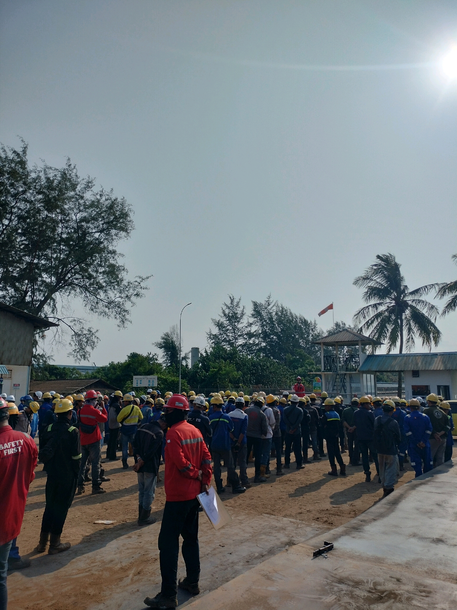 In Batam, Indonesia, a group of approximately 50 to 60 workers, dressed in safety vests and hard hats, are gathered outdoors in a dusty, industrial-like area. The weather appears sunny, with a bright sun visible in the upper right corner of the sky, casting long shadows on the ground. A few palm trees and other vegetation are present in the background, along with some low-rise buildings and a small tower structure with a flag. The workers are facing in the same direction, suggesting they are listening to a speaker or receiving instructions, though no speaker is clearly visible in the frame. Some individuals are wearing red or blue safety vests over their work clothes. The ground is a mixture of dirt and concrete.