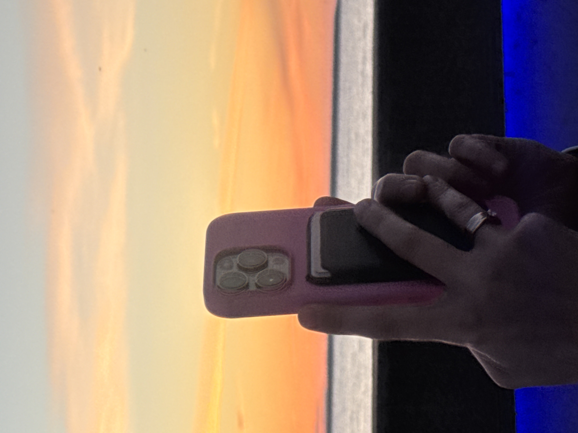 In the foreground, a person's hand is holding a pink-cased smartphone up to take a picture. The phone has a prominent camera module with multiple lenses. The person is wearing a ring on their finger. In the background, a body of water is visible, likely the Baltic Sea, with a thin strip of land or coast in the distance. Above the water, the sky displays a gradient of colors characteristic of a sunset or sunrise, with hues of orange, yellow, and pink. The overall scene suggests an outdoor setting, possibly a waterfront promenade or a balcony overlooking the sea. The specific location in Tallinn, Estonia, cannot be determined from the visual information alone, but the presence of the sea and the sky's colors are consistent with a coastal city at dusk or dawn. The lower part of the image shows a dark horizontal surface and a band of bright blue light, possibly artificial lighting.