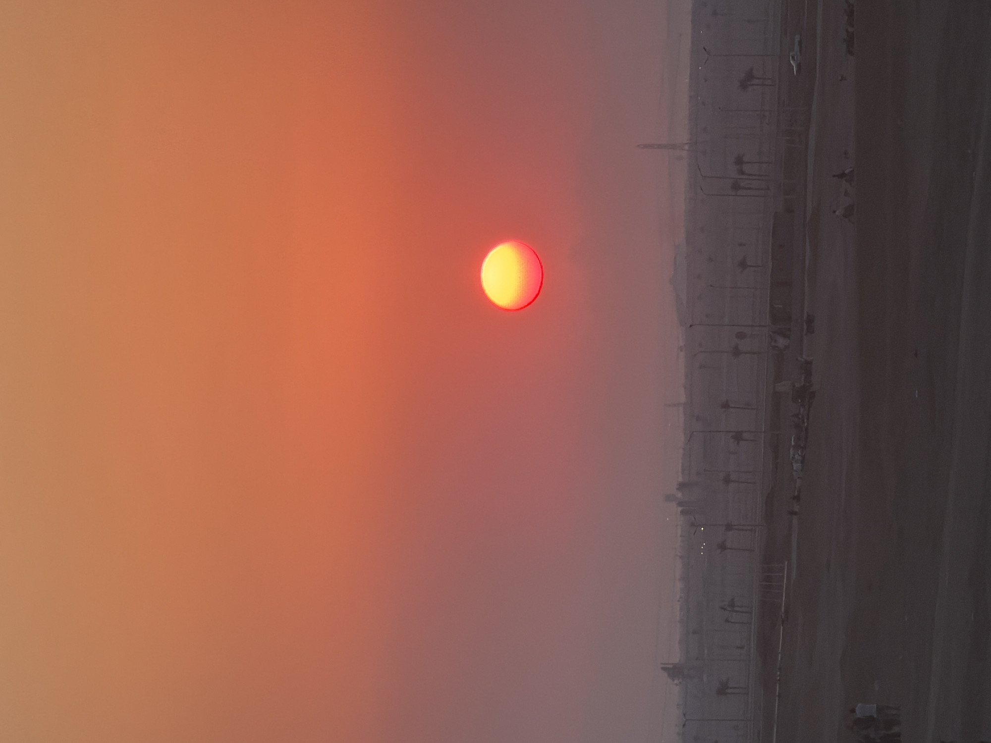 The image captures a sunset over a desert landscape, likely in or near Sharm El Sheikh, Egypt, given the presence of palm trees and construction. The sky is a vibrant gradient of orange and pink hues, with the sun appearing as a bright, reddish-yellow orb positioned slightly off-center in the upper right quadrant of the frame. In the mid-ground, a flat, dusty terrain stretches out, dotted with sparse vegetation, streetlights, and what appear to be the beginnings of urban development, including concrete structures and industrial elements in the distance. A few individuals are visible in the lower left and mid-ground, some standing in groups and others appearing to be engaged in activities like walking or sitting. A white vehicle is also present in the mid-ground. The overall atmosphere is hazy, contributing to the soft glow of the setting sun and the muted colors of the landscape.