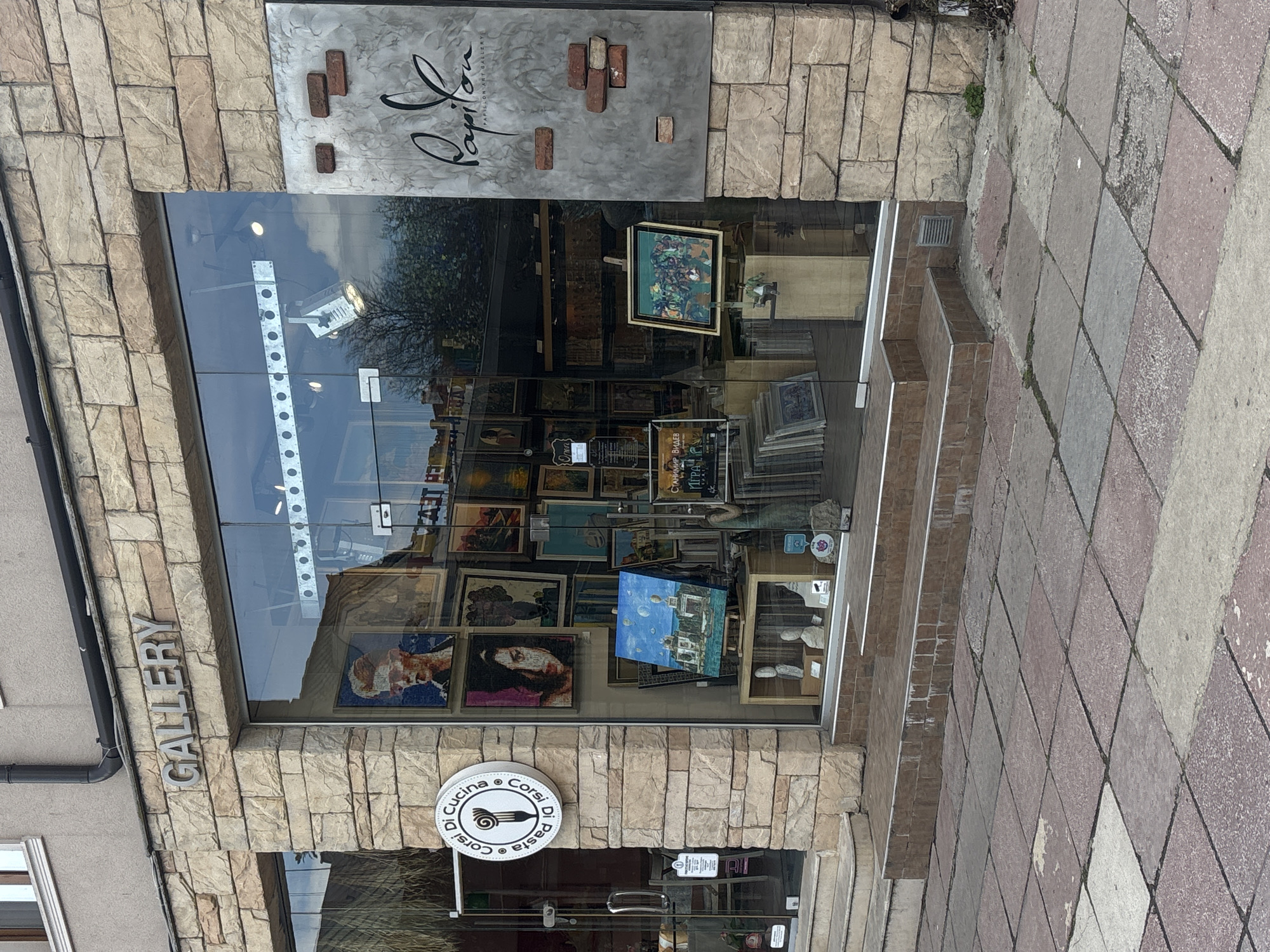 The image shows the exterior of an art gallery and pasta restaurant in Varna, Bulgaria. The gallery, named "Papilon Art Gallery," has a large glass window displaying numerous paintings and artwork. The building's facade is constructed from tan stone bricks. To the left of the gallery, the word "GALLERY" is vertically mounted on the wall. Below the gallery, the entrance to "La Cucina Corsi di Pasta e Corsi" is visible, marked by a circular sign depicting a fork and pasta. The sidewalk in front is paved with pink and gray diamond-patterned stones. Reflections can be seen in the gallery's window.