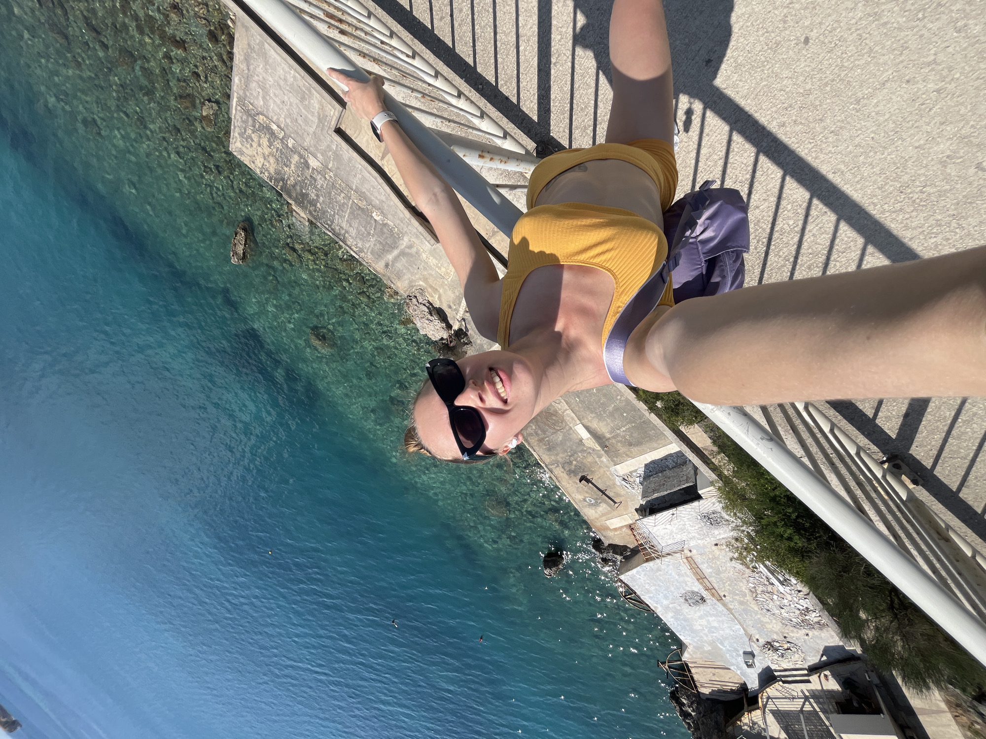 An inverted selfie captured from a high vantage point looks down over a serene body of water and concrete structures. The primary subject is a young woman, likely in her 20s or early 30s, smiling broadly at the camera. She wears black sunglasses, a yellow ribbed crop top, and yellow shorts. A white smartwatch adorns her left wrist, and a purple bag or backpack rests beside her. Her light-colored hair is pulled back, and she appears to be holding onto a white metal railing, which casts long, distinct shadows across the light-colored concrete path she stands on.
Below the woman and the railing, the clear, blue-green water reveals rocks and the seabed, indicative of a shallow, possibly coastal or lakeside environment. Adjacent to the water are various concrete platforms and structures, some appearing somewhat dilapidated or under construction, with debris or broken pieces scattered on them. Lush green vegetation grows along the edge of these structures near the water.
The activity depicted is a casual selfie, taken during what appears to be a sunny day with clear skies, suggested by the bright light and sharp shadows. The woman's joyful expression conveys a relaxed and pleasant mood. There is no visible text in the image.