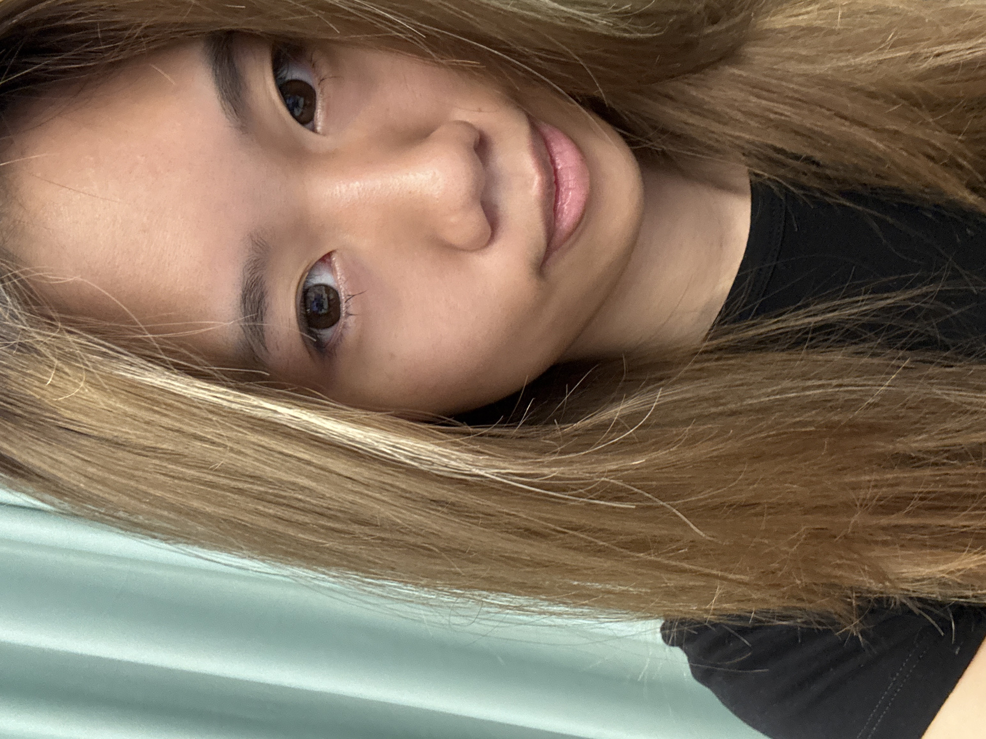 The image depicts a close-up selfie of a young woman with light brown hair. She has brown eyes and a fair complexion. She is wearing a black shirt. The background includes a light blue or green surface. The photo does not contain enough details to confirm the location in Nan'an District, China.