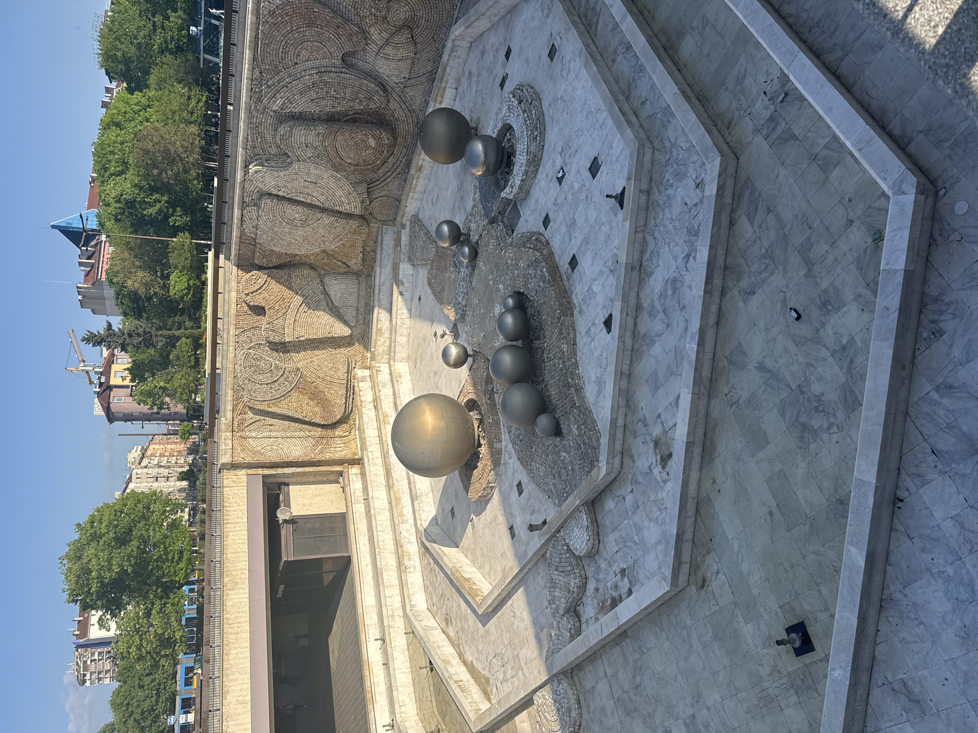 The image shows the Cascade, a monument located in Sofia, Bulgaria. The monument features large decorative walls with mosaic patterns and a series of metal spheres arranged in a tiered fountain structure. The fountain is dry. The ground surrounding the monument is paved with marble tiles. Trees and buildings are visible in the background, indicating an urban setting. There are some objects such as trash.