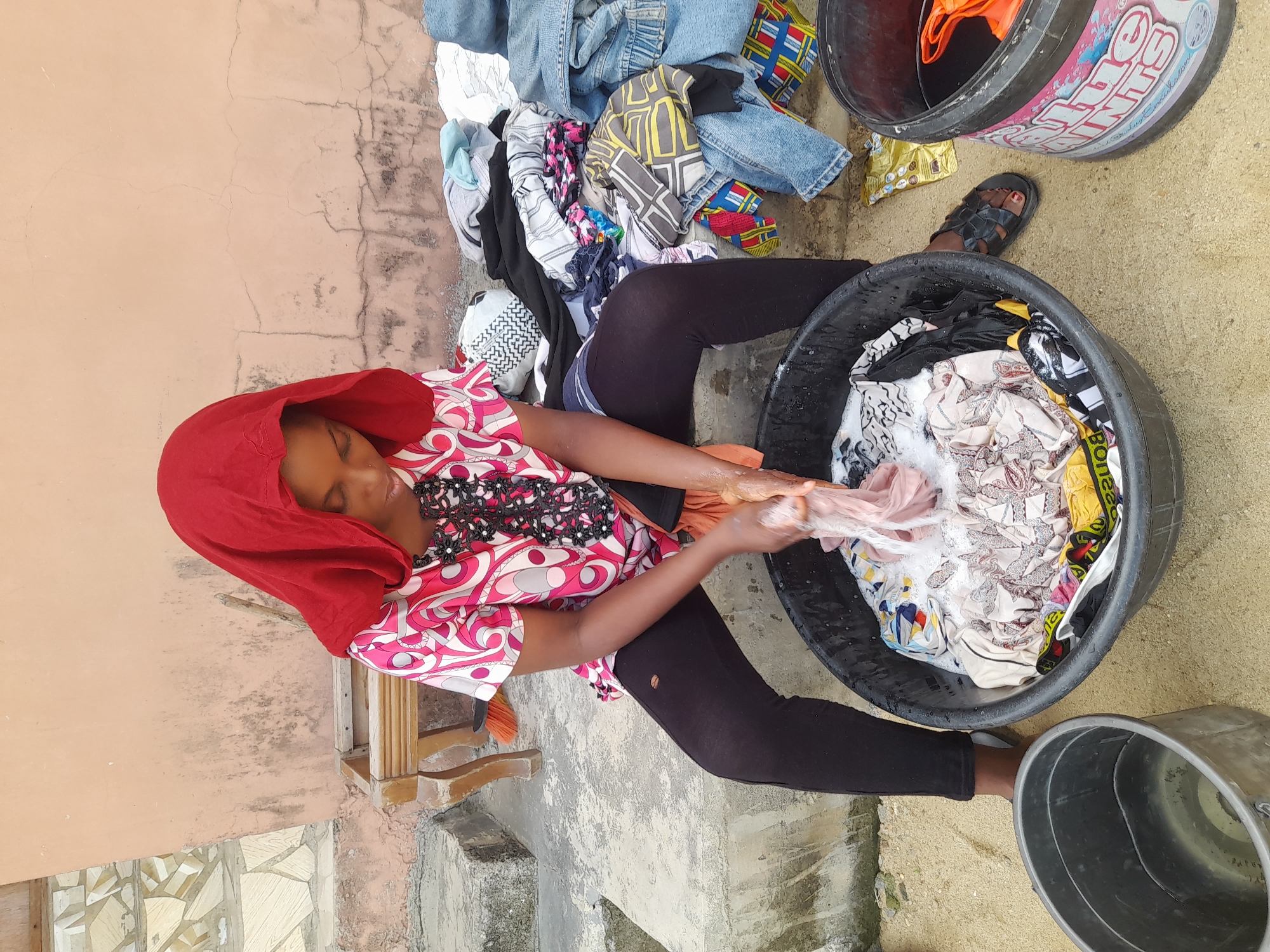 A person is depicted handwashing clothes outdoors. The individual, who appears to be a woman, is seated on the ground with her legs slightly bent, facing a large black plastic basin filled with clothes and soapy water. She wears a vibrant red head covering that frames her face, a colorful patterned top with pink, white, and black designs, and dark leggings. Her arms are submerged in the basin, actively scrubbing a light-colored garment.
To her left, a large pile of various clothes, including denim jeans, patterned fabrics, and light-colored items, is stacked on the ground. Another black bucket, partially visible behind the main basin, contains more clothes, one of which has "Bona" written on it. To the right, another metal bucket is present, bearing the faded text "Shine Paints" in a distinctive pink and white logo. A pair of black sandals is seen near this bucket.
The setting appears to be an outdoor area with a concrete or tiled ground that has patches of dirt and moisture. A light brown wall, showing cracks and some discoloration, forms the background. The lighting suggests it is daytime. The overall scene depicts a common chore of laundry being performed manually in a utilitarian outdoor environment in Ado, Nigeria.