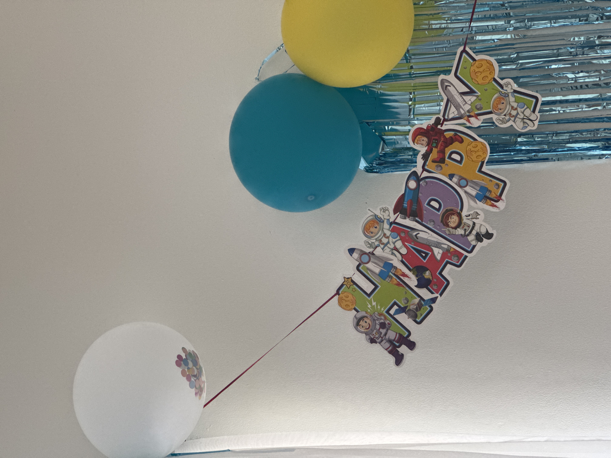 The image shows a decorated ceiling with a yellow and a blue balloon at the top, a metallic blue streamer, and a string banner that says "Happy 4th." The banner features astronaut and space-themed cartoon images on each letter. A white balloon with a cluster of small, colorful circles is at the bottom left, connected to the banner by a red string. The background is a plain white ceiling. The location is not discernible from the image.