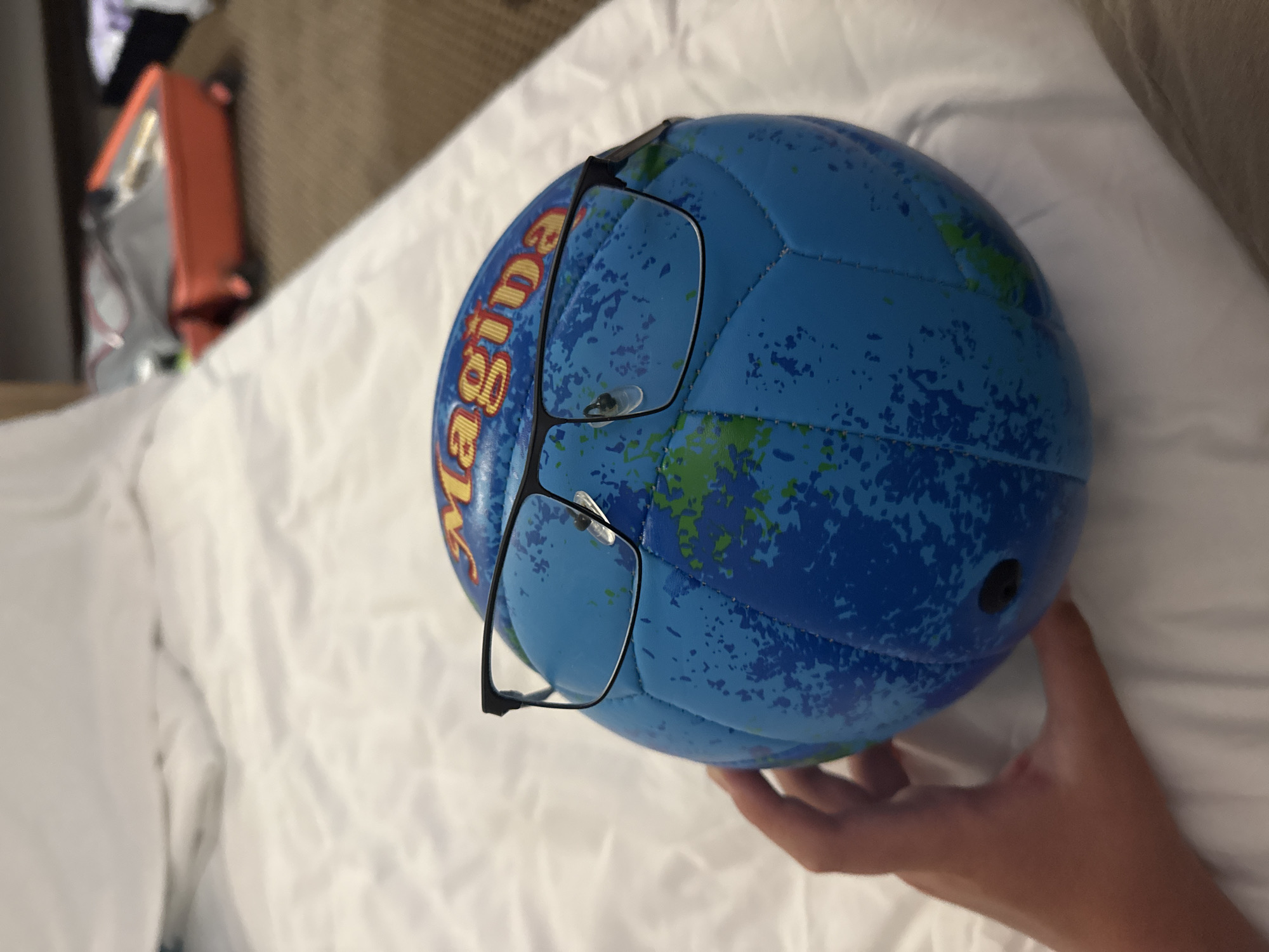 A hand holds a blue and green globe-patterned volleyball that has been fitted with black-rimmed glasses. The ball is positioned on a white, textured surface, possibly a bed. In the blurred background, an orange suitcase is visible, along with other luggage. The image does not provide any information or context related to the city of Nesebar, Bulgaria.