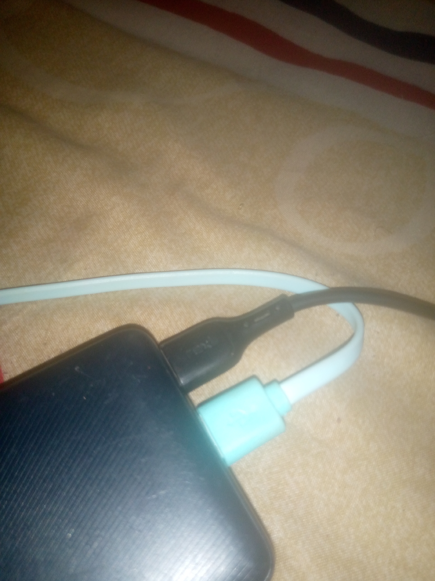 The image shows a close-up of a dark blue power bank and two charging cables, one black and one light blue, plugged into it. The objects are resting on a textured, light brown fabric surface with visible red and dark stripes in the upper portion of the image. The lighting is dim, casting shadows and highlighting the textures of the fabric and the power bank. There are no people or identifiable locations in the image.