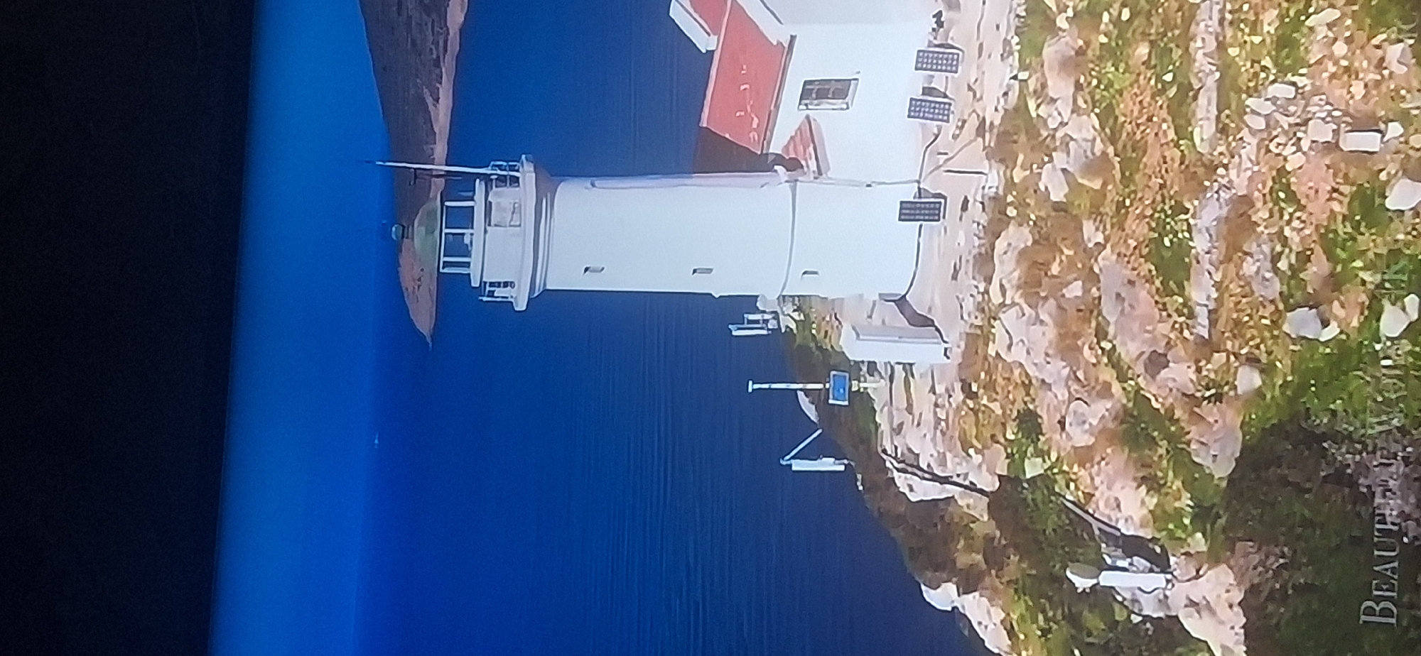 The image shows a white lighthouse with a red-roofed building attached, situated on a rocky cliff overlooking a deep blue body of water. The lighthouse has a green-domed top and a lantern room with windows. Several antennas and solar panels are visible on the cliff near the lighthouse. The landscape surrounding the lighthouse is rocky and sparse, with some patches of green vegetation. In the distance, a hilly landmass can be seen. A faint text overlay at the bottom of the image reads "BEAUTIFUL WORLDS". The location is not in the city of Bila Tserkva, Ukraine, as the scene depicts a coastal environment with a lighthouse, which is not characteristic of Bila Tserkva.
