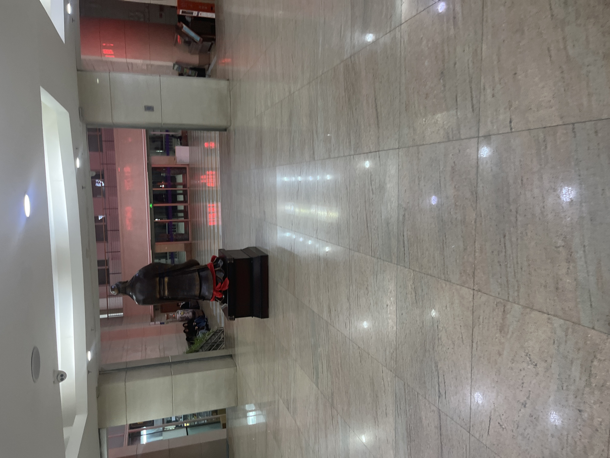 The image depicts the interior of what appears to be a public building, possibly a transportation hub, in Changchun City, China. The floor is covered in large, light-colored stone tiles. A bronze statue, adorned with a red sash, stands on a dark platform in the center of the frame. In the background, there are entrances and waiting areas. People are visible in the background, likely waiting or passing through the area. There is also a red sign visible. The location is likely in Changchun City, China.