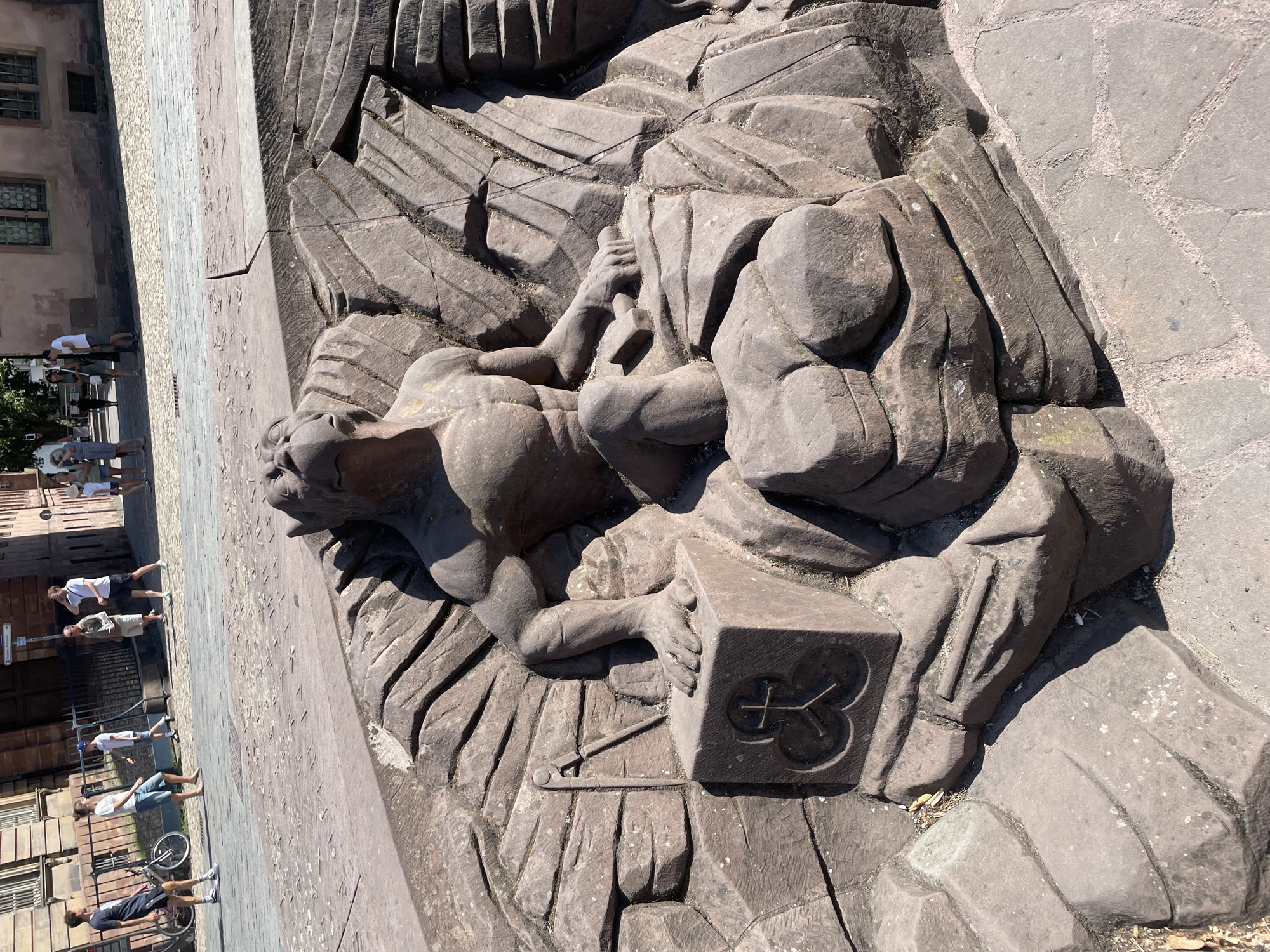 This image captures a stone sculpture in a public square in Strasbourg, France. The sculpture depicts a seated figure with a lion-like head, muscular human torso, and avian wings or features. The figure is surrounded by rough-hewn stone formations resembling rocks or geological strata. In its left hand, the figure grasps a stone cube bearing a symbolic emblem. To the left of the figure, a surveying tool, possibly a compass and straightedge, is carved into the stone. The sculpture appears to be part of a fountain or decorative installation. In the background, people are visible walking on a cobblestone street, with historic buildings lining the square. The overall scene is bathed in sunlight, suggesting a daytime setting.