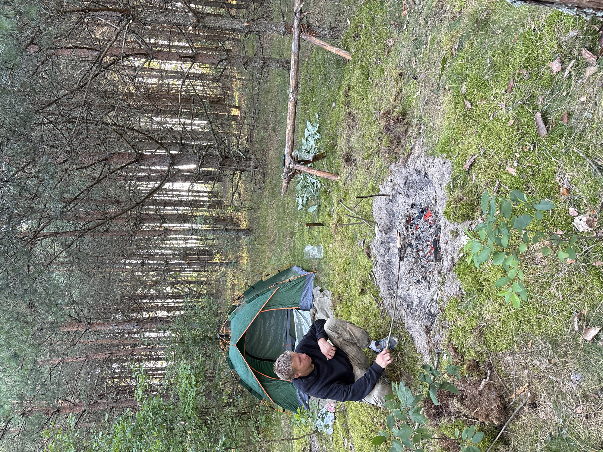 A young man with blond hair is crouching in a forest clearing near a green tent. He is wearing a black hoodie and camouflage pants. He is holding a stick over a campfire that is smoldering with red embers. In the background, there is a wooden structure with some leaves piled next to it. The ground is covered in moss and pine needles. The forest appears to be dense with tall pine trees. The scene suggests a camping trip in a rural area of Belarus.