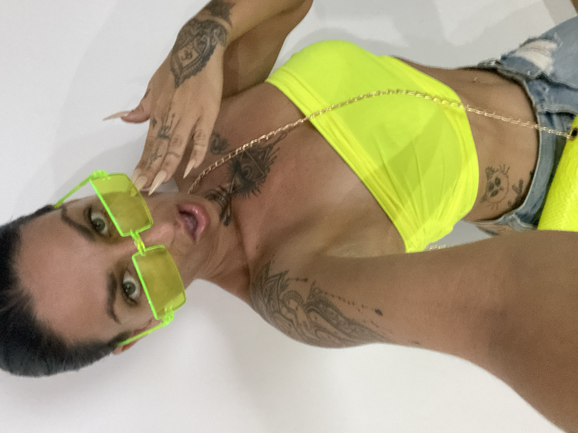 A woman with dark hair pulled back and green eyes is wearing bright yellow rectangular sunglasses. She has several tattoos, including one on her chest and others on her arms and hands. She is also wearing a neon yellow strapless top and ripped denim shorts, with a gold chain strap across her chest. The background is a plain white wall. The image appears to have been taken indoors. There is no information in the image that would indicate the location is Mũi Né, Vietnam.