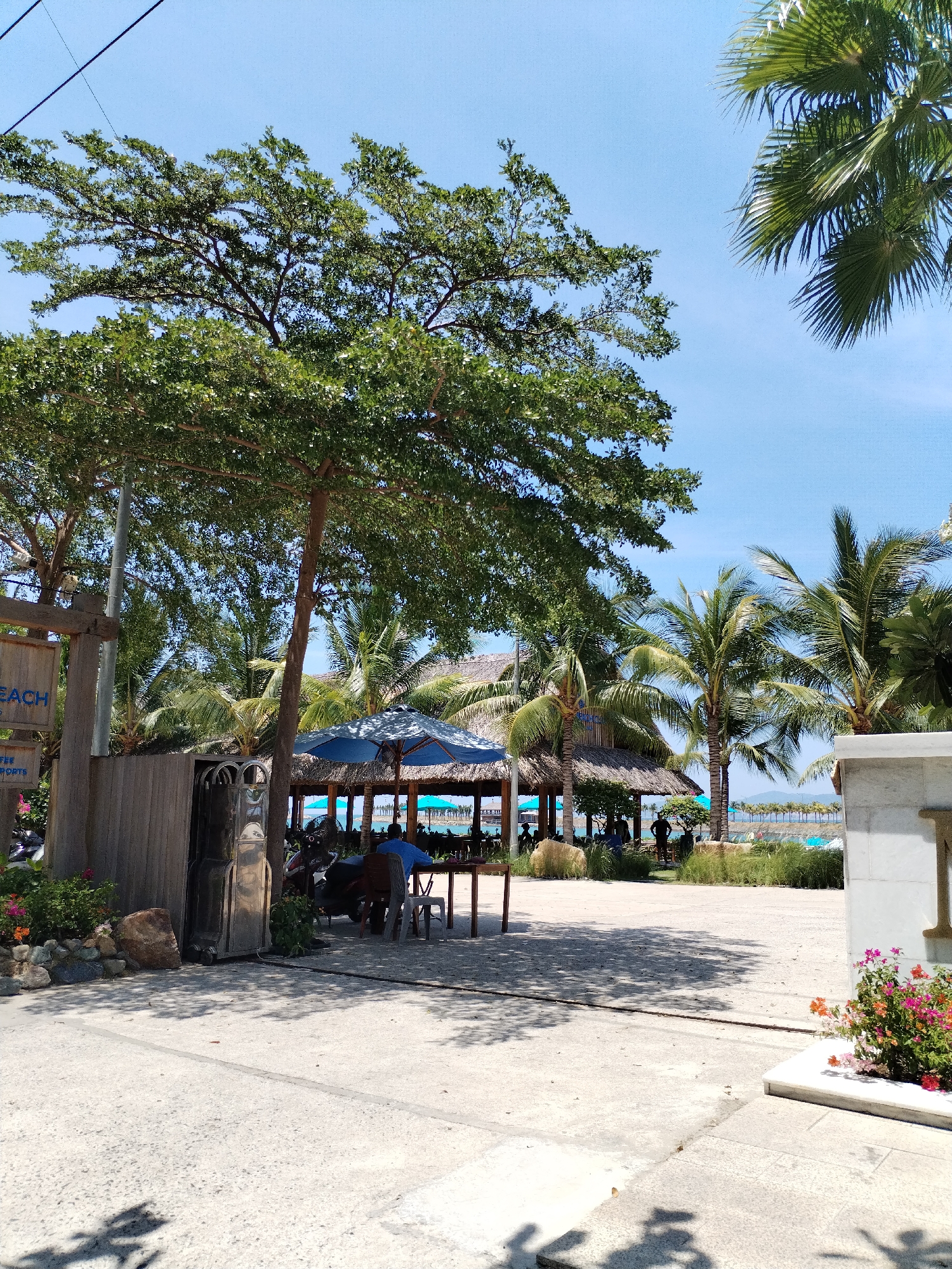 The image depicts an outdoor scene in Bac Nha Trang Ward, Vietnam, with a paved foreground transitioning into a sandy area that leads to a beach. Several palm trees and other lush vegetation are visible, framing a thatched-roof structure in the middle ground. Underneath a blue parasol are visible chairs and table on the sand. In the distance, the sea and beach are dotted with further parasols and people. A weathered wooden sign is to the left, and a white structure with flowers is on the right. There are shadows cast by trees across the paved area.