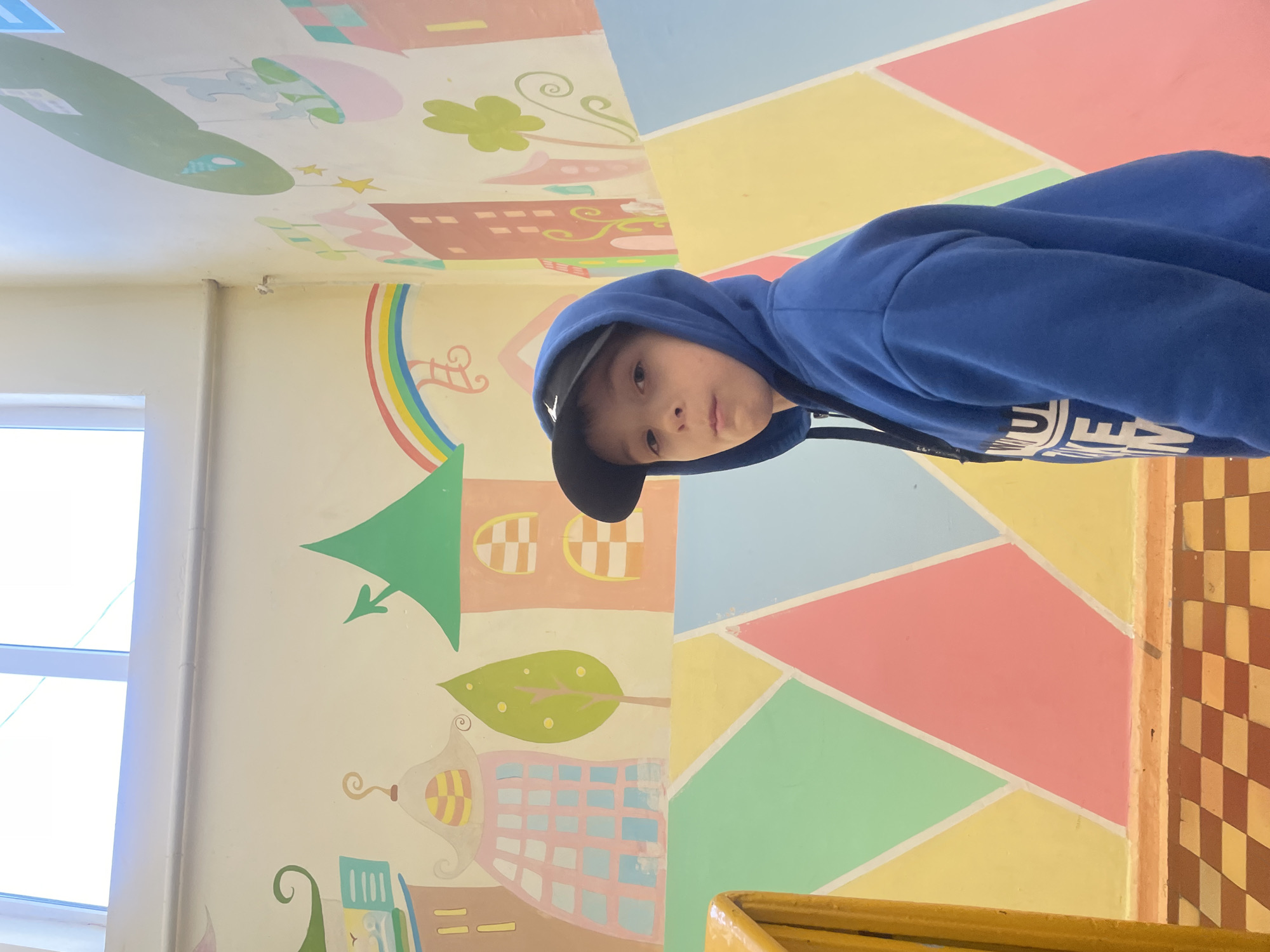 A young boy is depicted indoors, looking directly at the camera with a neutral expression. He wears a bright blue hooded sweatshirt, with the hood up, and a black baseball cap worn backward. Partially visible white text on his hoodie includes the letter "U" followed by other indistinct letters.
The setting appears to be a child-friendly environment, possibly a classroom, daycare, or play area in Sochi, Russia. The walls and possibly the ceiling are elaborately decorated. One wall features whimsical, cartoon-like illustrations of houses, a green castle, a rainbow, leaves, stars, and abstract swirls, rendered in pastel shades of cream, light green, pink, yellow, and light blue. Another section, likely an adjacent wall or ceiling, displays a geometric pattern of large, irregular polygons in similar soft colors such as yellow, pink, light blue, and mint green.
On the far left, a large white-framed window allows bright natural light into the room, indicating it is daytime. In the bottom right corner, a section of a checkered floor in brown and lighter brown/yellow tones is visible, alongside a yellow-colored railing or low barrier. The boy appears to be posing for the photo.
