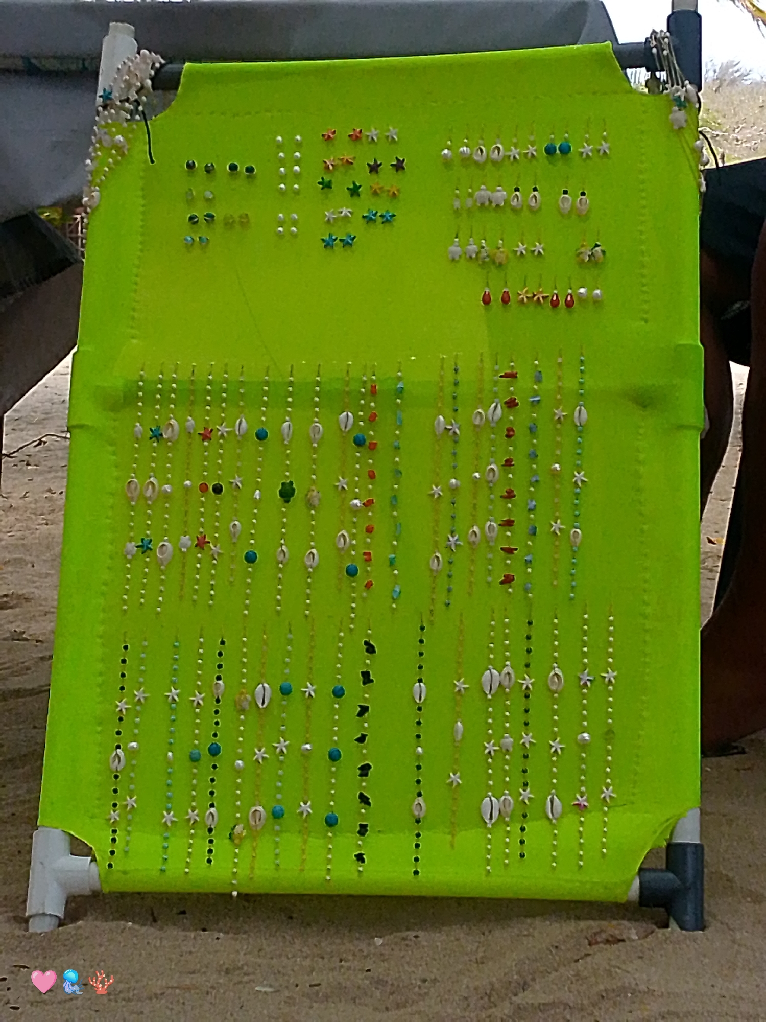On a beach in Pedro González, Venezuela, a light green display stand with jewelry is set up on the sand. The jewelry includes earrings with beads and starfish and beaded strands with shells and starfish. The stand has a white leg on the front left and a gray leg on the front right. The beach sand is light brown, and the lower-left corner of the image contains heart, whale, and coral emojis.