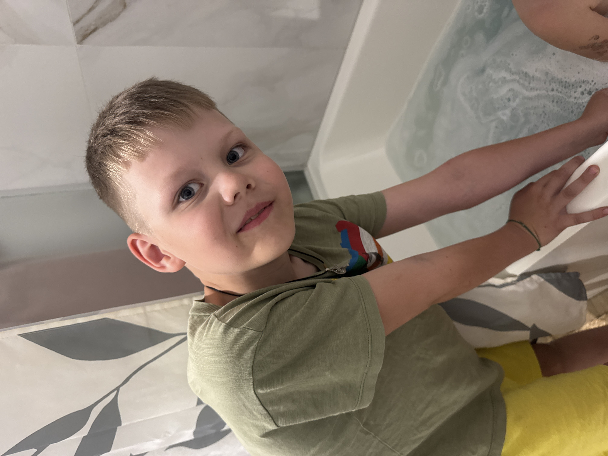 A young boy with short, blond hair and blue eyes looks up at the camera, a playful expression on his face. He is wearing a light green t-shirt with a colorful patch on the chest and a thin black necklace. The boy is in a bathroom, indicated by the tiled walls and a bathtub filled with bubbly water. He is leaning over the edge of the tub, his right arm resting on the rim, and his left arm reaching towards the water. His skin appears fair and he has a few faint freckles across his nose. The lighting suggests it is daytime.