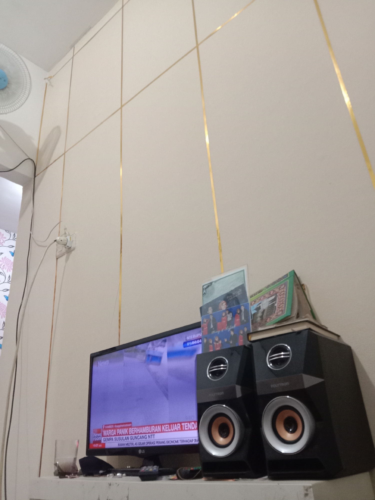 The image depicts an indoor scene, likely a living room or entertainment area, in Brangsong, Indonesia. The dominant feature is a television screen displaying a news broadcast with Indonesian text. To the right of the television are two black speakers, stacked one on top of the other. On top of the speakers, there are several items, including what appear to be books and a framed picture or certificate. A wall with decorative gold lines runs across the background. A ceiling fan is partially visible in the upper left corner. Electrical outlets and cables are also present on the wall. The overall setting suggests a domestic environment.