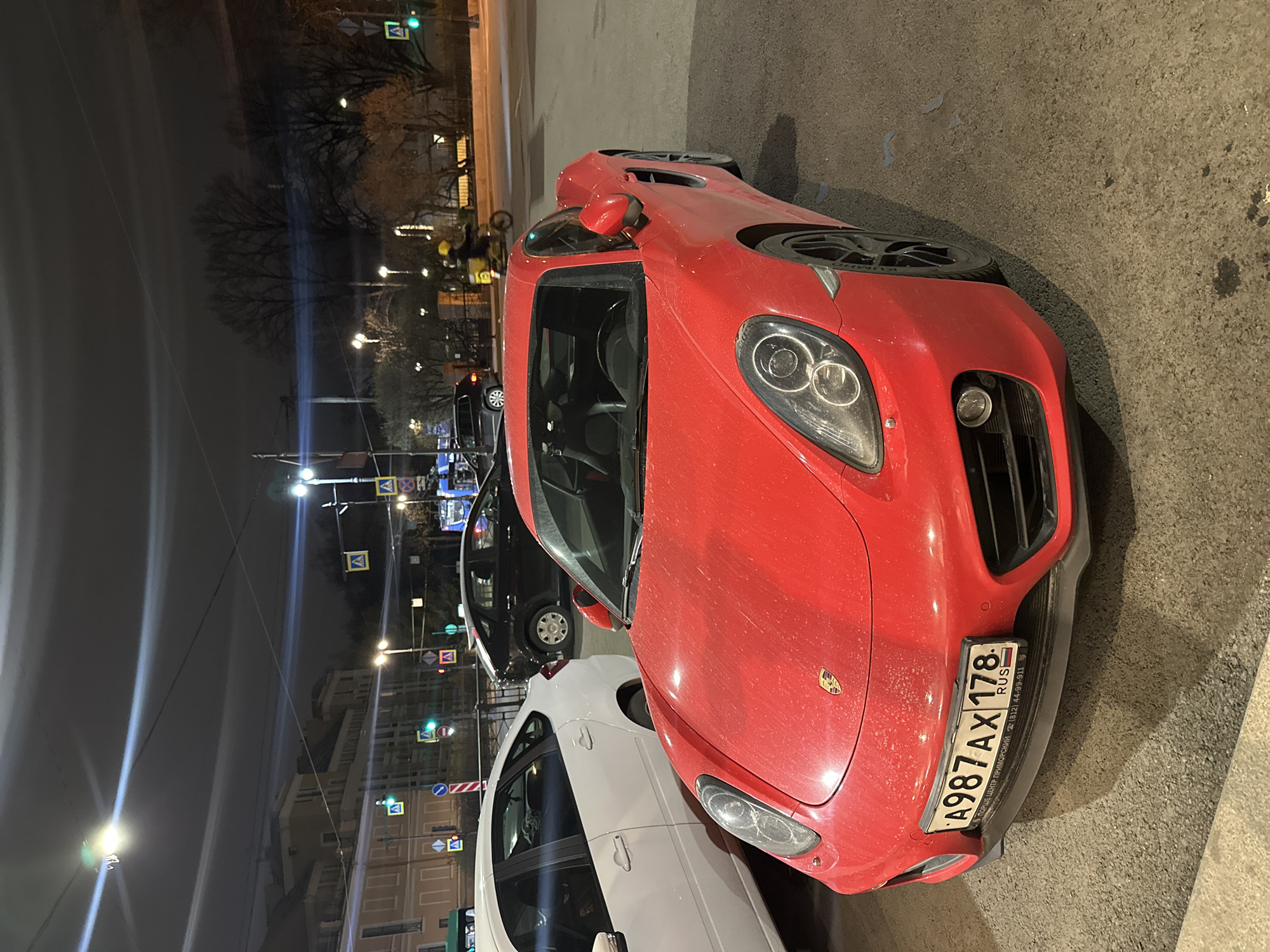 The image captures a street scene at night in Saint Petersburg, Russia. Dominating the foreground is a red Porsche Cayman, parked at an angle on a paved surface. The car's license plate, which is partially obscured, reads "A987AX 178 RUS." Behind the Porsche is a white car, partially visible, and a black car further away. In the background, street lights illuminate a street lined with buildings and various road signs. The scene indicates an urban environment with parked vehicles and street furniture. There is no human presence in the visible frame.