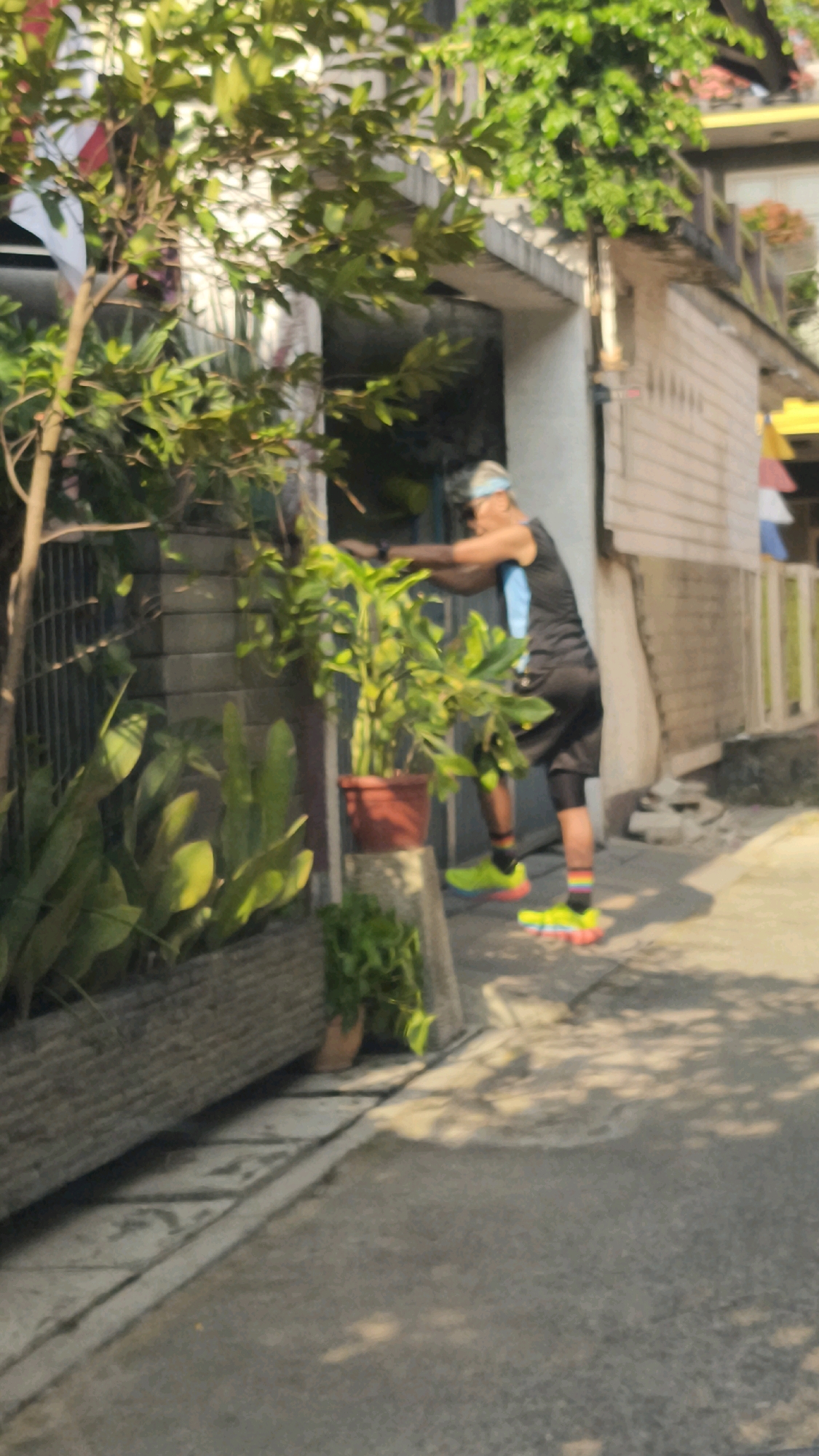 The image depicts a person engaged in physical activity, possibly stretching or exercising, in an outdoor urban environment. The person, wearing a dark tank top, black shorts, and brightly colored running shoes, is leaning forward with their arms extended. They appear to be in front of a building with white horizontal siding and a dark entrance. Lush green plants, including a potted plant and a planter box filled with foliage, are present on the left side of the scene, creating a natural element amidst the urban setting. The ground is paved with concrete, and sunlight casts dappled shadows, suggesting it is daytime. The background shows more of the building and a fence. Based on the provided information, this scene is located in the city of Bekasi, Indonesia.