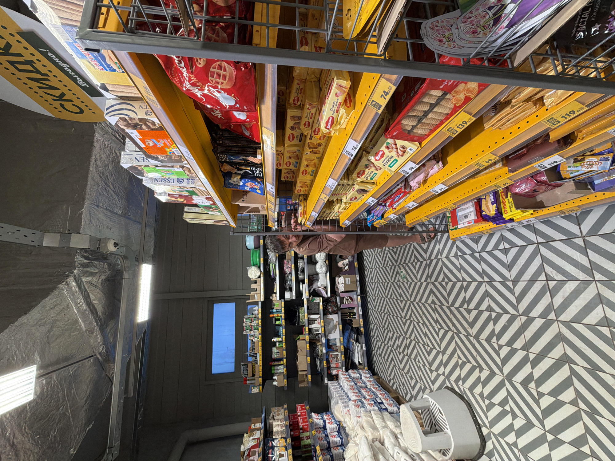 Here is a description of the image:
The image shows the interior of a store, presumably a supermarket or a similar retail environment, with shelves full of various products. The focus is on shelves stocked with snacks, candies, and baked goods such as cookies and waffles, which are neatly arranged with price tags visible on the shelves.
A person is visible standing near the shelves, seemingly looking at the products. The aisles of the store contain other shelves stocked with various items, including groceries and household supplies. The floor has a distinctive patterned tile design, with alternating white and gray tiles creating a geometric effect. The store appears to be well-lit, with lighting fixtures on the ceiling. The image likely originates from Chaikovsky, Russia, given the presence of Russian brand names and price labels.