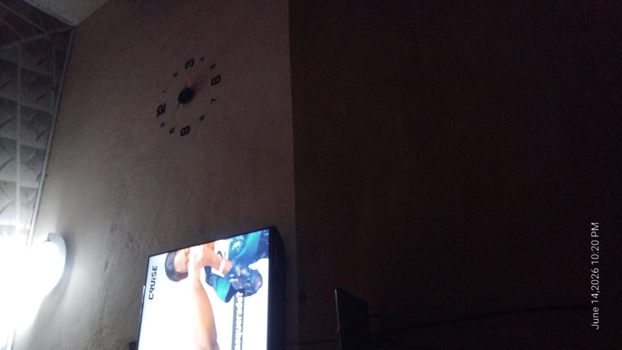 The image shows a wall with a clock and a television. The clock has numbers 1 through 12 arranged in a circle, and its hands are pointing to approximately 2:55. The television screen displays an image with the word "CRUISE" at the top. A person's arm is visible in the television screen, and the text "asquerad?" is visible below it. A light fixture is mounted on the ceiling above the television. The bottom of the image shows the date "June 14, 2026 10:20 PM". The scene appears to be indoors, likely a living room or entertainment area. The lighting is dim, with the television screen and ceiling light providing most of the illumination.