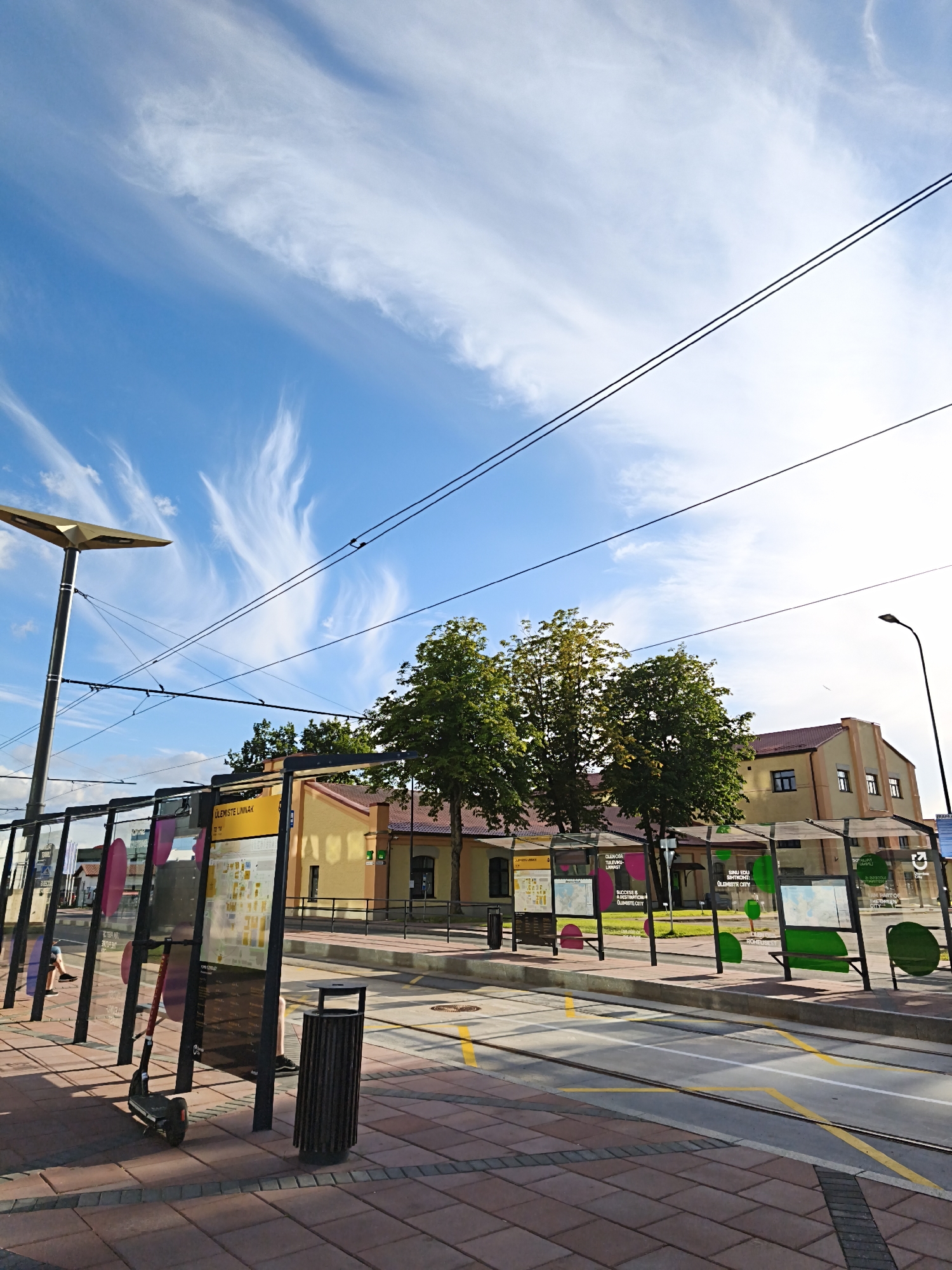 The image depicts a street scene in Tallinn, Estonia, specifically at a tram stop. The sky is a vibrant blue with wispy white clouds, and overhead, tram wires are visible against the sky. In the foreground, a modern tram stop features glass-enclosed shelters with informational displays. An electric scooter is parked near the shelter, and a trash bin stands next to it. The ground is paved with reddish-brown bricks. Behind the tram stop, a yellow building with a red roof is visible, flanked by trees. A street lamp with a distinctive modern design is also present on the left side of the image. In the distance, another street lamp and a portion of a larger building are seen. The overall impression is of a well-maintained urban public transport area on a sunny day.