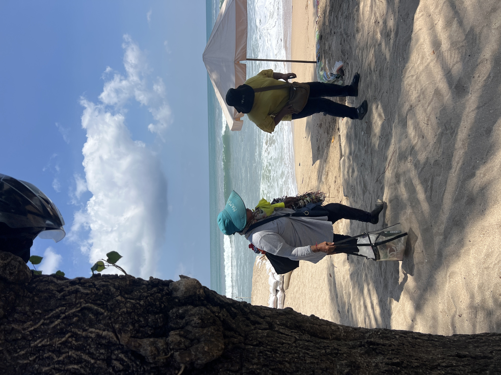 Two women stand on a sandy beach in Kuta, Indonesia. The woman on the left wears a blue hat, a grey long-sleeved shirt, black pants, and dark shoes. She carries a black bag over her shoulder and a transparent bag in her right hand. The woman on the right wears a black hat, a yellow short-sleeved shirt, black pants, and dark sandals. She carries a brown bag across her body. Behind them, the ocean has moderate waves, and a white beach umbrella stands on the sand. Sandbags are visible in the background. The scene is lit by daylight under a partly cloudy sky. A tree trunk occupies the left foreground.