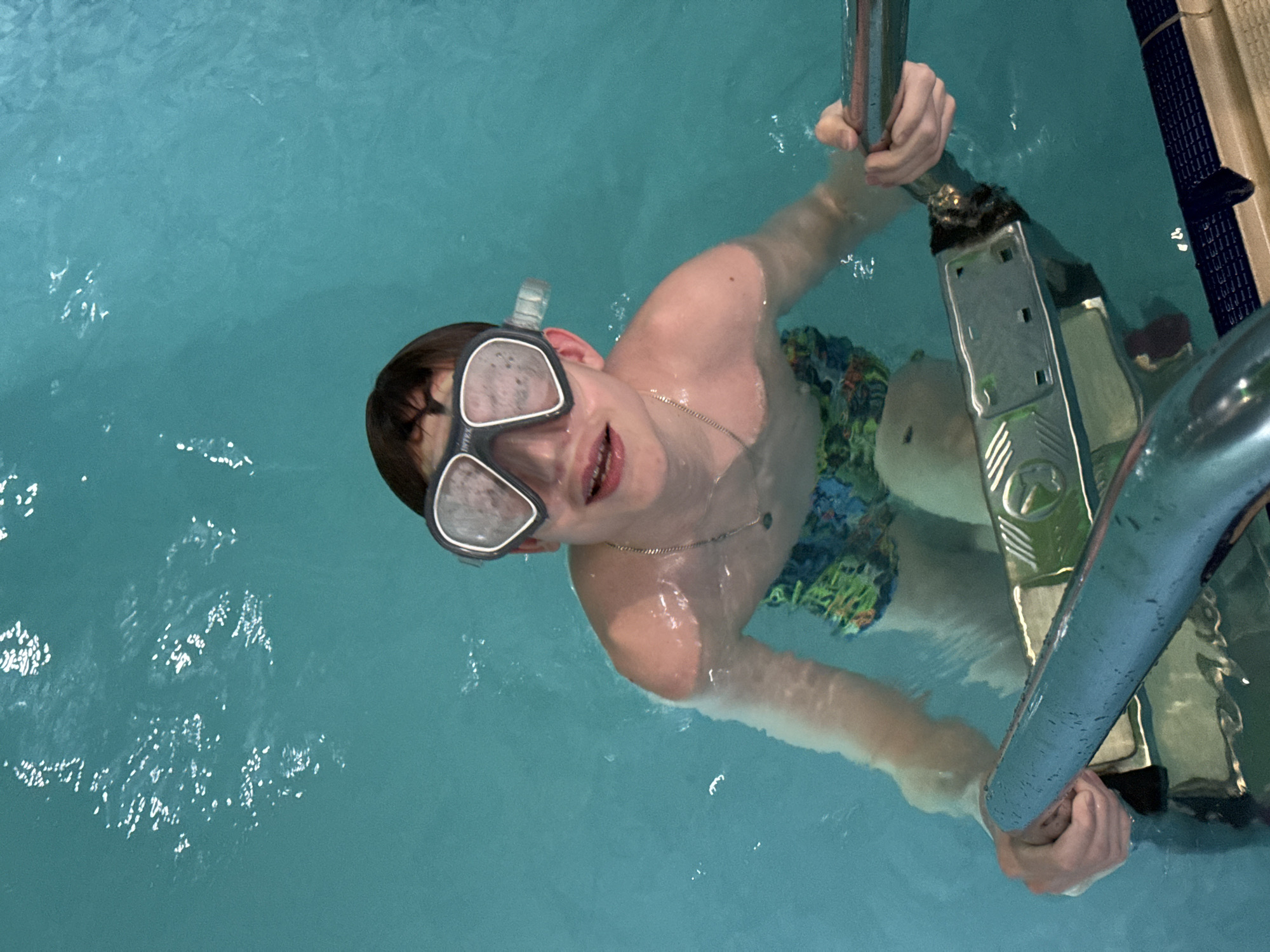 A fair-skinned young male is in a swimming pool, wearing swimming goggles and patterned swim trunks. He is holding onto a metal ladder positioned along the edge of the pool. The pool water is a clear turquoise color. It's not possible to determine the exact location in the city of Chaikovsky, Russia, from the image.