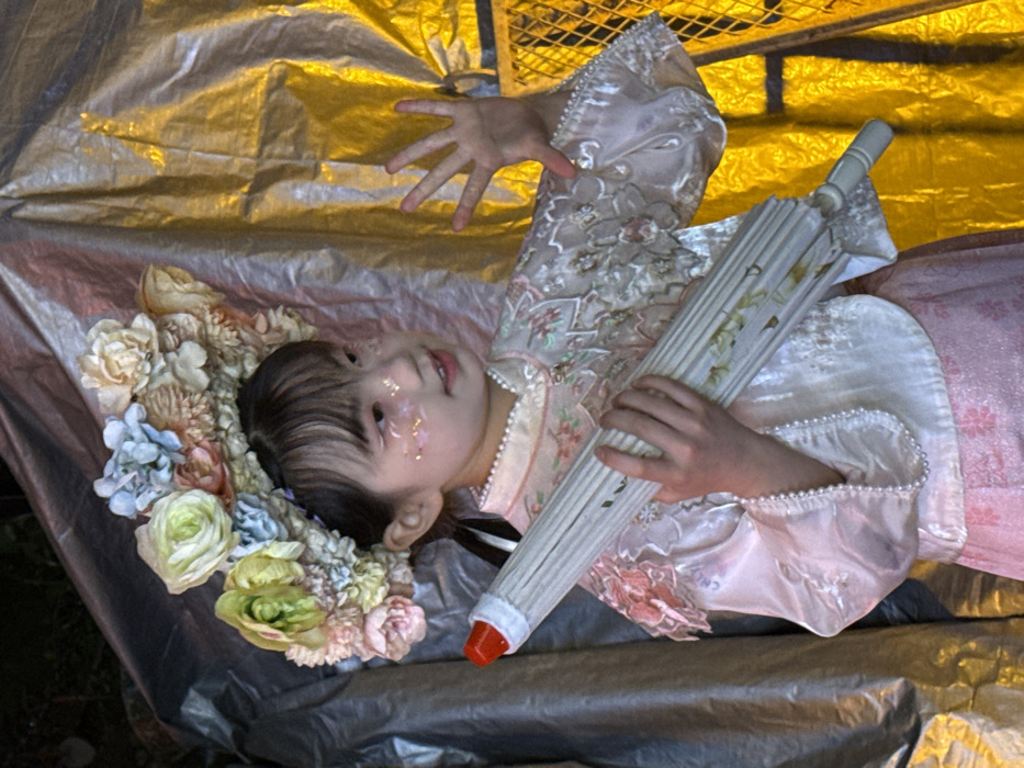 A young person, adorned with a floral crown and glitter on their face, lies on a reflective surface under golden lighting. They wear a light-colored, floral-patterned garment with pearl trim and hold a closed umbrella with a red tip. One hand is extended, fingers spread open, toward the top of the frame. The scene suggests a fairytale or whimsical theme.