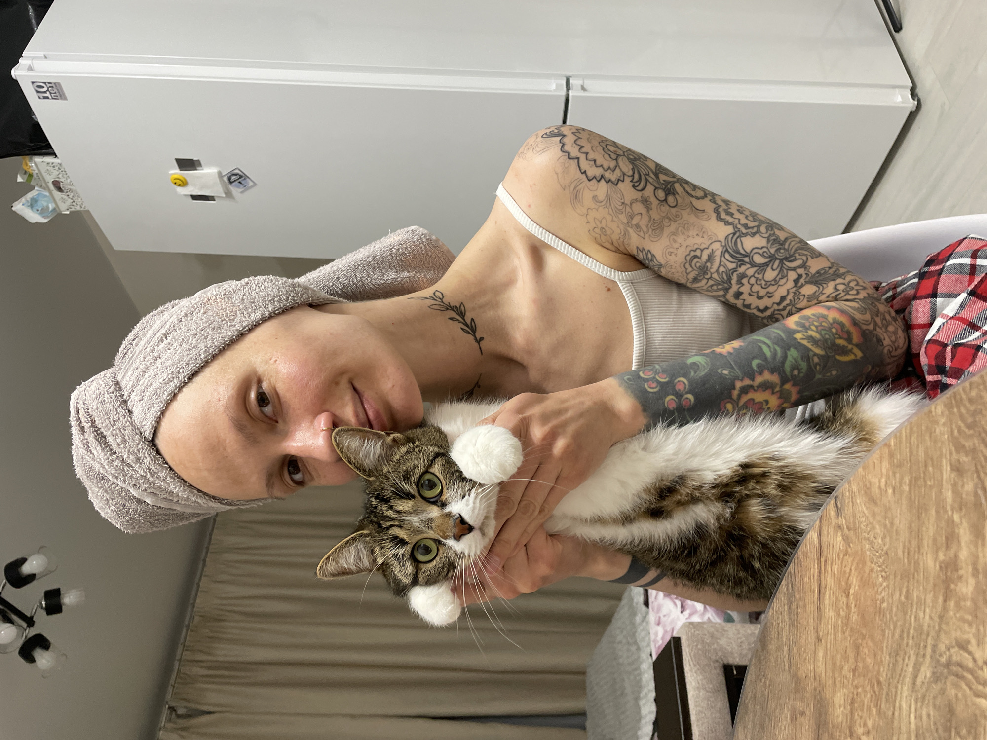 A woman with extensive tattoos and a towel wrapped around her head sits indoors, holding a cat. She is wearing a white tank top, with the strap visible on her left shoulder, and a plaid red and black garment partially visible behind her right arm. Her left arm and shoulder are adorned with intricate floral and mandala-style tattoos, while a delicate leafy vine tattoo is on the right side of her neck. She has a subtle nose ring and is looking directly at the camera with a gentle expression.
The cat, a tabby and white with bright green eyes and prominent white paws, is being cradled in the woman's hands, looking towards the viewer. Its fur is a mix of brown, black, and white patches.
The background shows a white refrigerator on the left with a "10 In" label and a few magnets, including a yellow smiley face. On the right, part of a wooden table surface is visible. Above the woman's head, a dark light fixture with multiple bulbs can be seen, and a beige curtain hangs in the background. The lighting suggests an indoor setting during the daytime or a well-lit evening. The overall scene conveys a sense of warmth and affection between the woman and her pet.