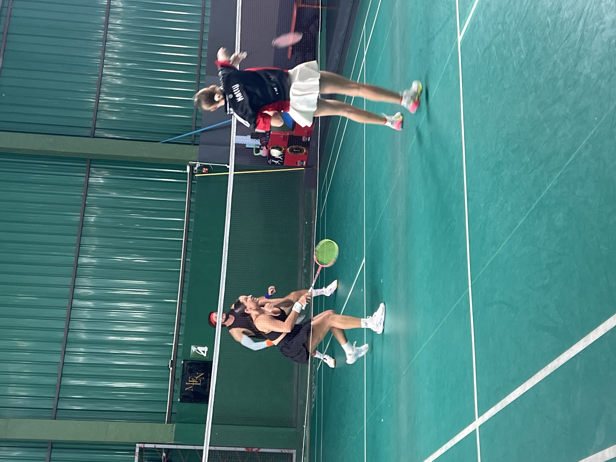 In a badminton court in Ban Rang Rak, Thailand, four individuals are engaged in a doubles match. The court is illuminated, with the green floor marked by white lines. The walls are covered with green corrugated metal. In the foreground, a woman in a black athletic outfit and a white skirt is in motion, holding a badminton racket with a yellow and pink head. Her opponent, a woman in a black and red shirt with "Kathy" printed on the back and a white skirt, is also in motion, hitting a shuttlecock. In the background, two male players are on the court. One, wearing a black tank top and a red headband, is positioned behind the net. The other male player is mostly obscured from view. Equipment, including weights and a yellow pole, are visible against the back wall. The lighting suggests an indoor setting.