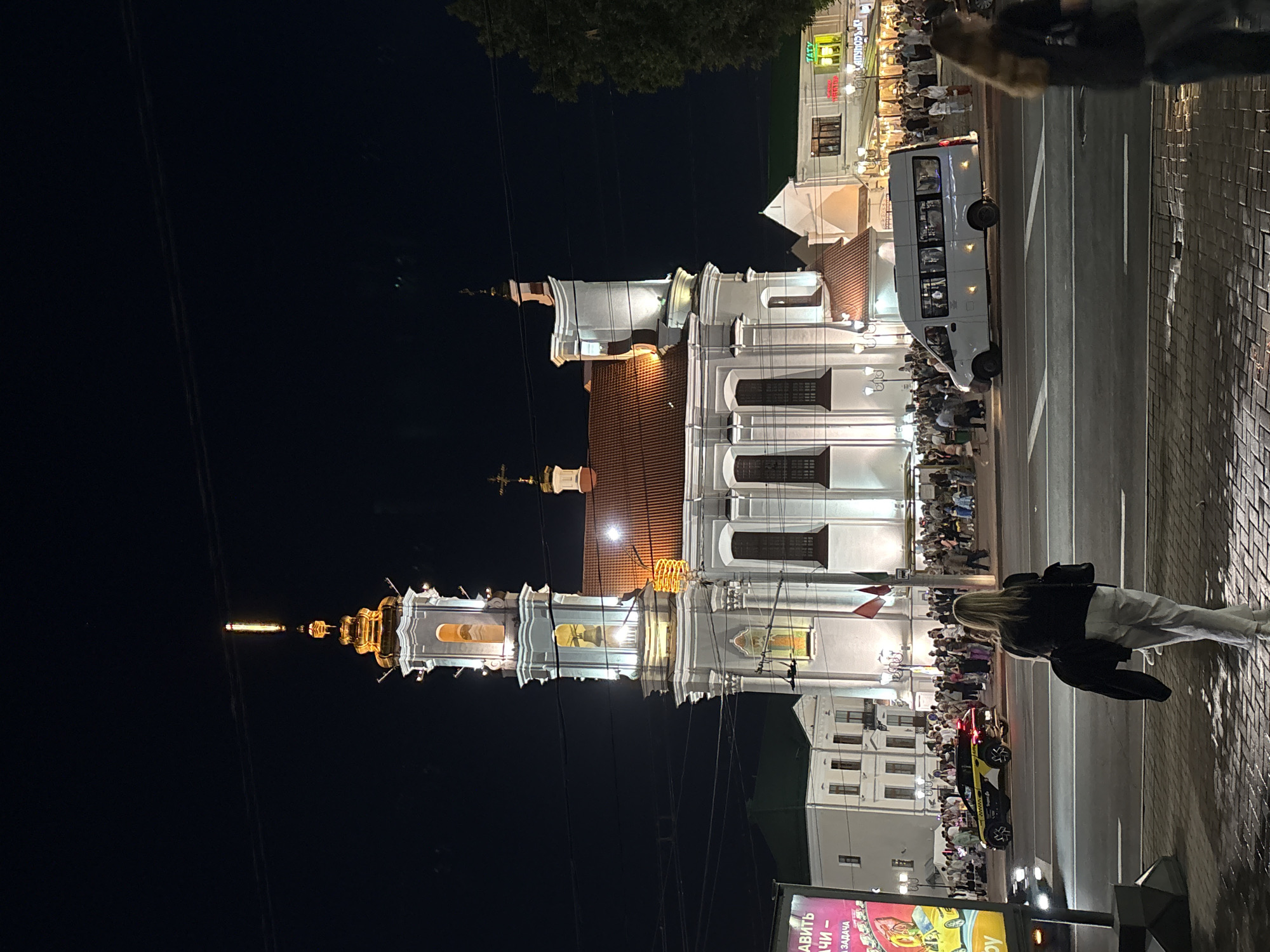 This is a night-time, eye-level, full-frame shot of the Holy Assumption Cathedral in Vitsebsk, Belarus. The cathedral is illuminated and stands prominently against a dark sky. In the foreground, the back of a woman with blonde hair wearing a black top and light-colored pants is visible, facing away from the camera and towards the street. A sidewalk made of cobblestones is beneath her feet. Across the street, a crowd of people is gathered in front of the cathedral. A white passenger van is driving on the street, and a yellow and black taxi is parked nearby. Several buildings with illuminated windows and signs are visible in the background, to the left and right of the cathedral, suggesting a commercial area. Power lines are visible crisscrossing the sky. A large billboard is on the left side of the frame. The overall scene depicts a lively urban environment at night, with a focus on the illuminated cathedral and the people in its vicinity.